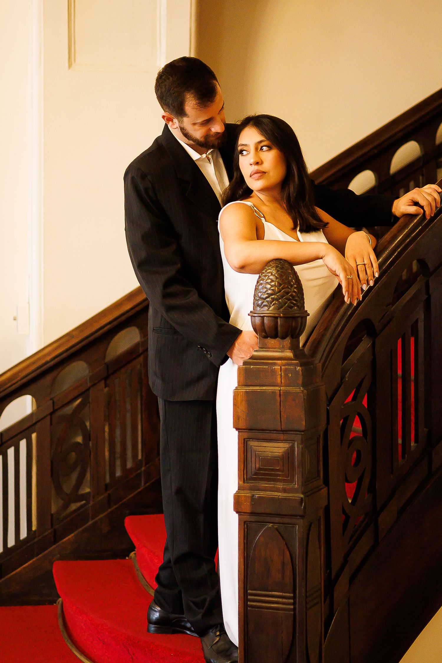Elegant couple in formal attire posing on a grand wooden staircase with red carpet, man in black suit, woman in white gown.