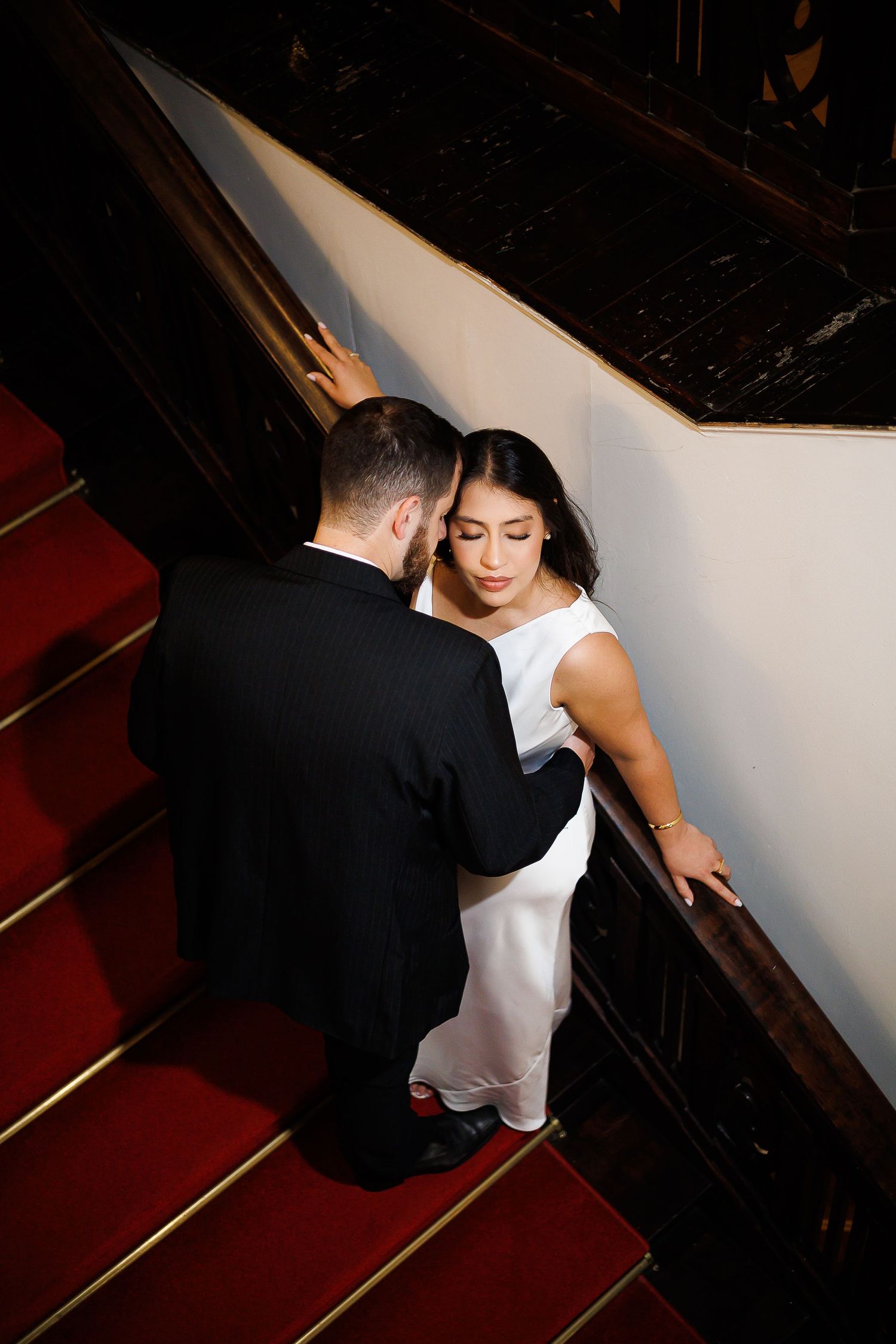 Bride in white gown and groom in black suit share an intimate moment on red-carpeted stairs with dark wood railings.