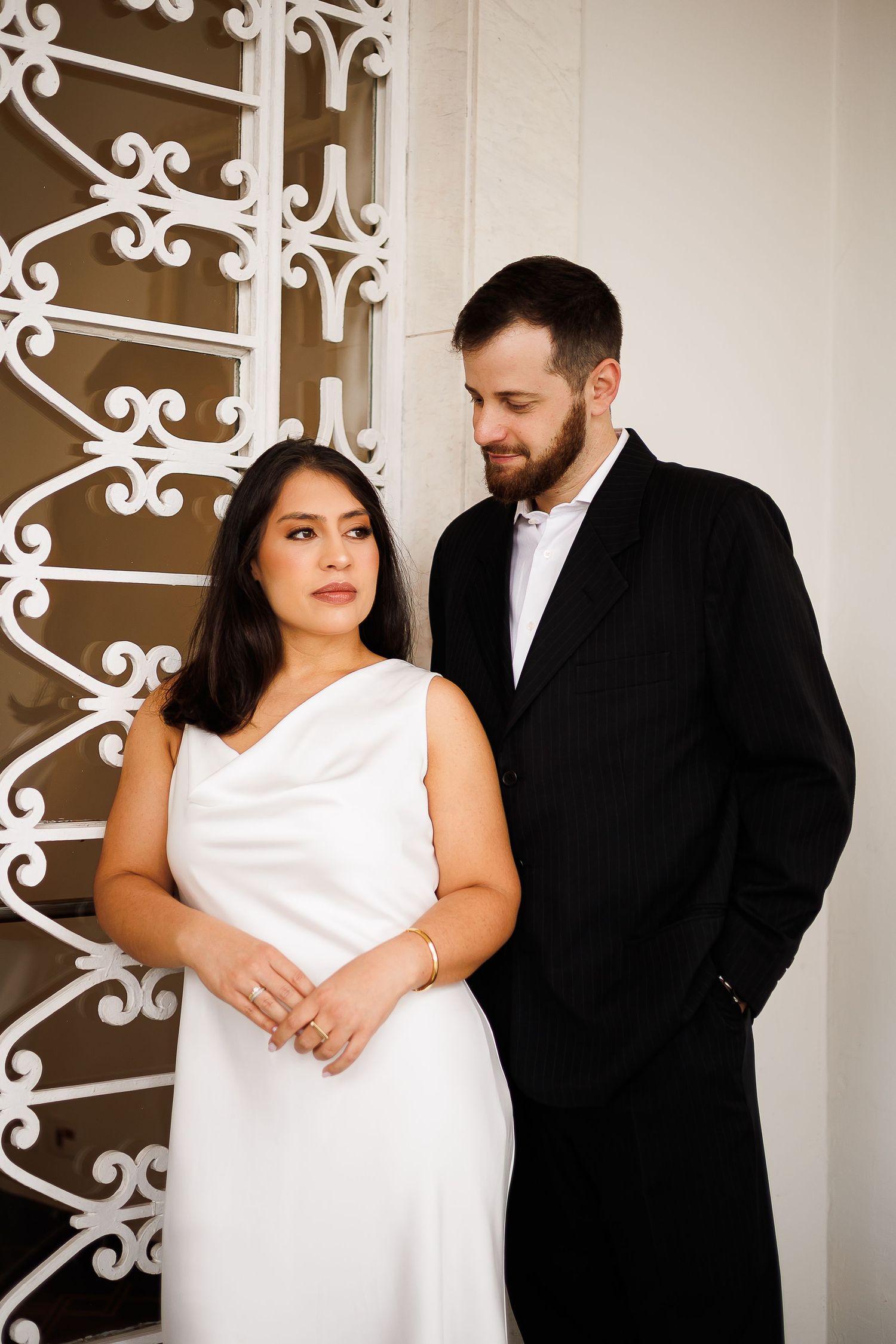 Elegant couple in wedding attire pose by ornate white ironwork, bride in white gown, groom in black suit.