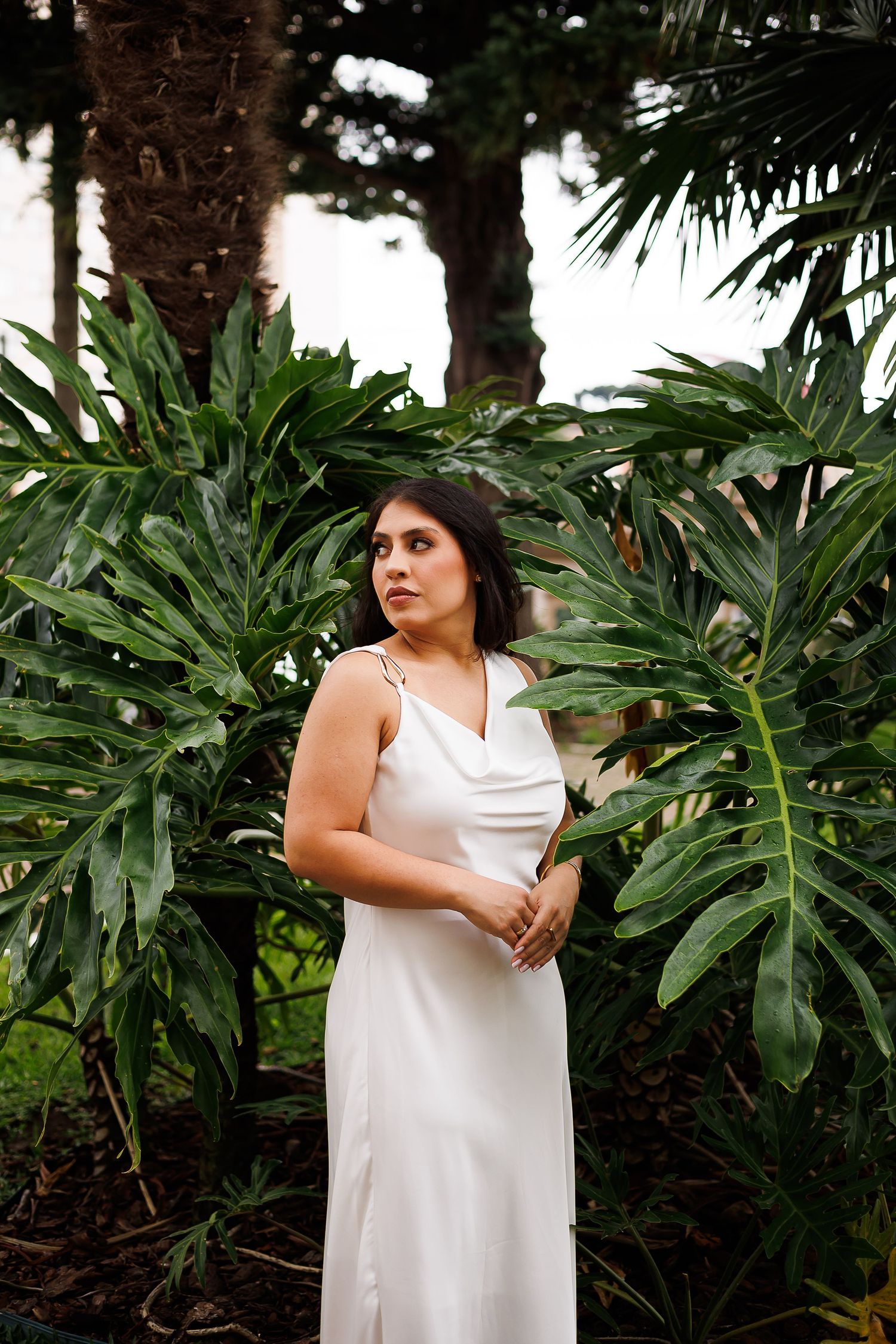 A bride in a white satin slip dress stands among lush tropical green plants and palm trees outdoors.