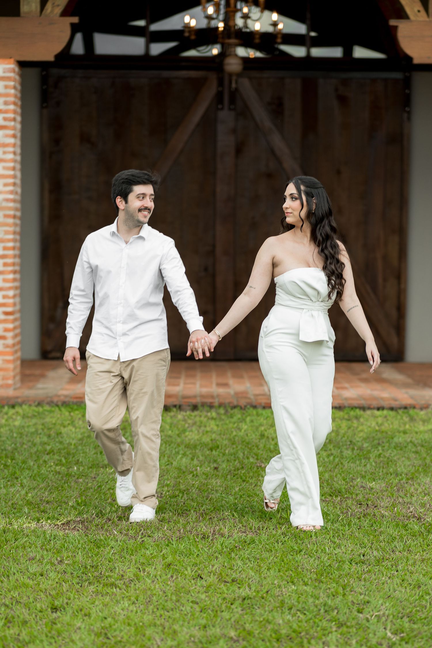 Smiling couple in white outfits holding hands strolling on lawn before wooden barn doors with string lights.
