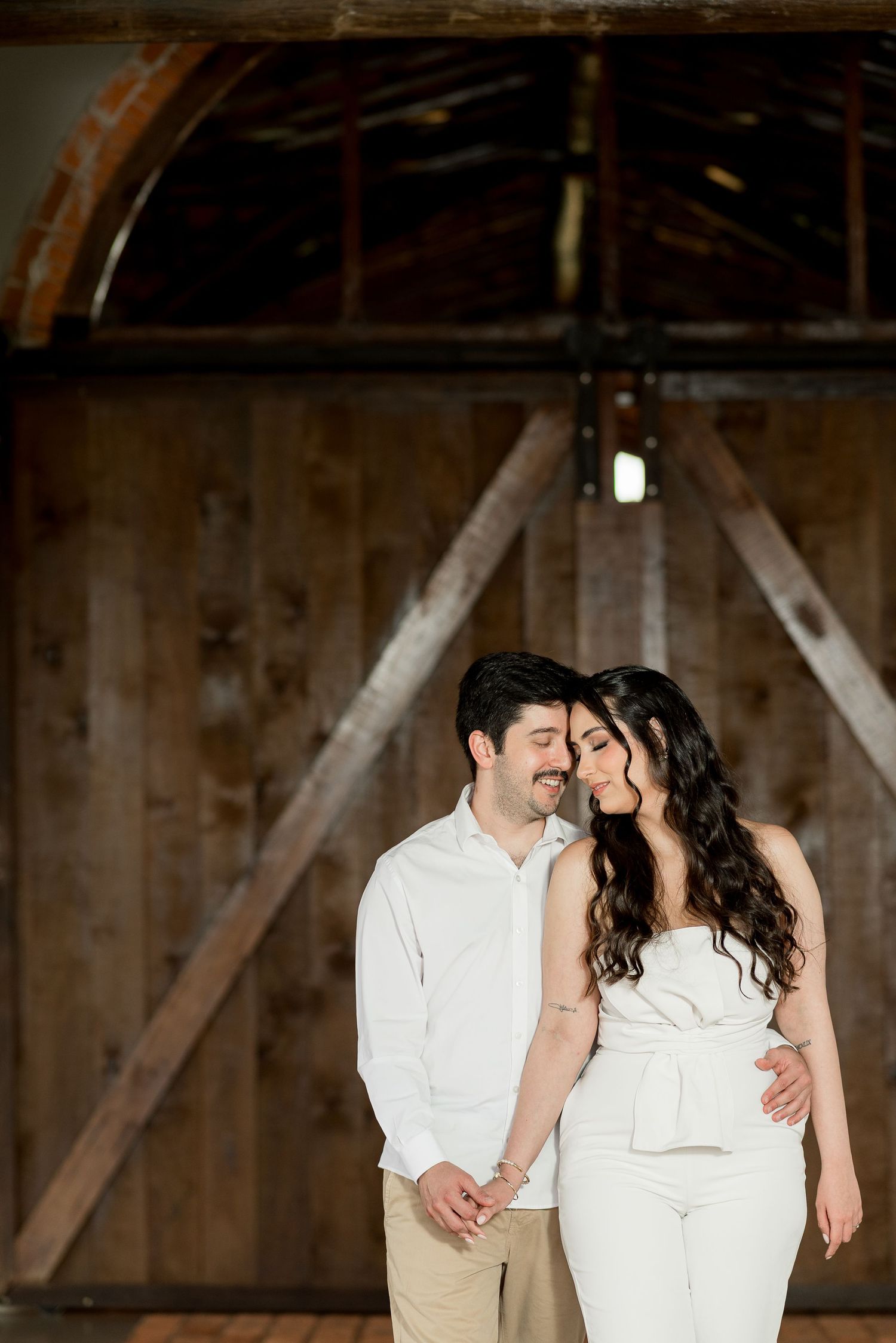 A couple in white outfits share a tender moment in front of rustic wooden barn doors during an engagement photo session.