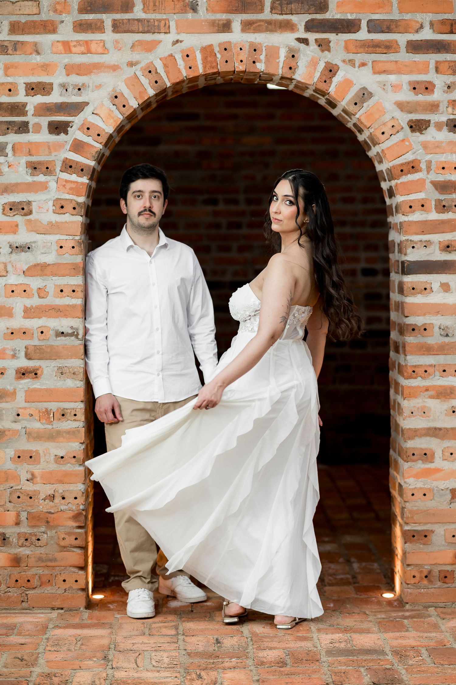 Couple posing in brick archway, bride in flowing white strapless gown, groom in white shirt and khaki pants.