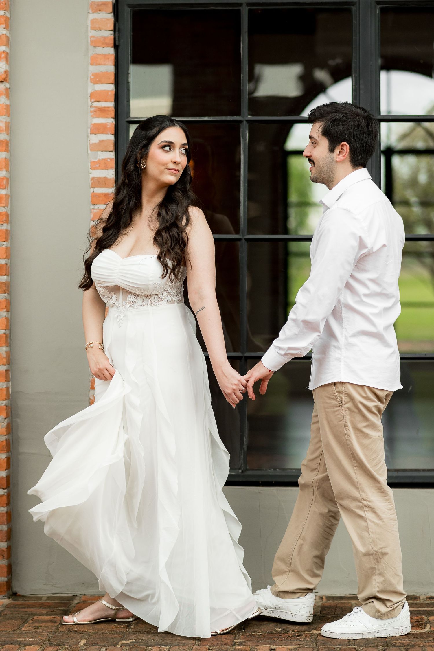 Bride in white gown holds hands with groom in white shirt and khakis outside brick building.