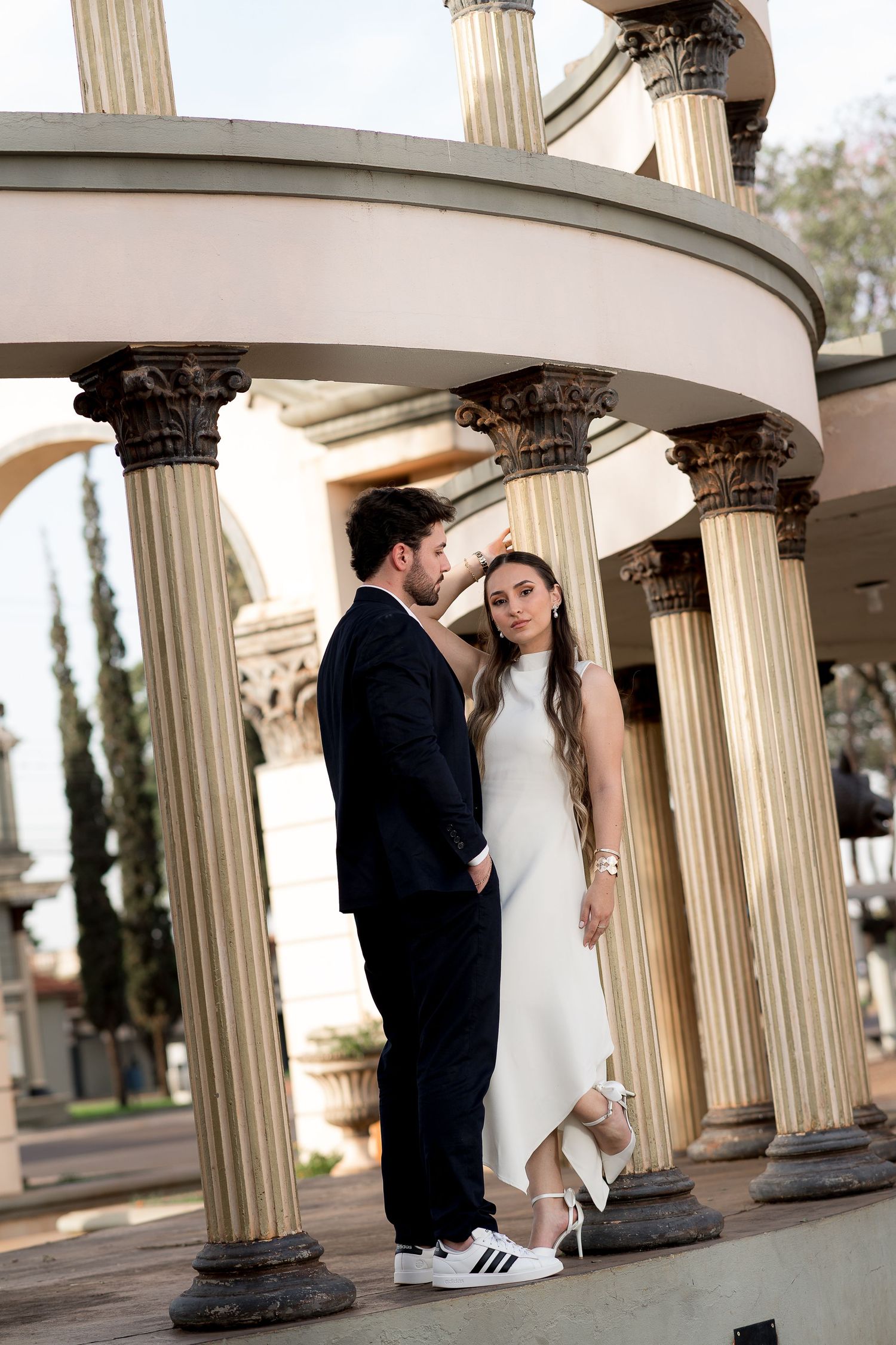 Couple in formal attire poses among classical columns, man in dark suit touches woman's hair in white dress.