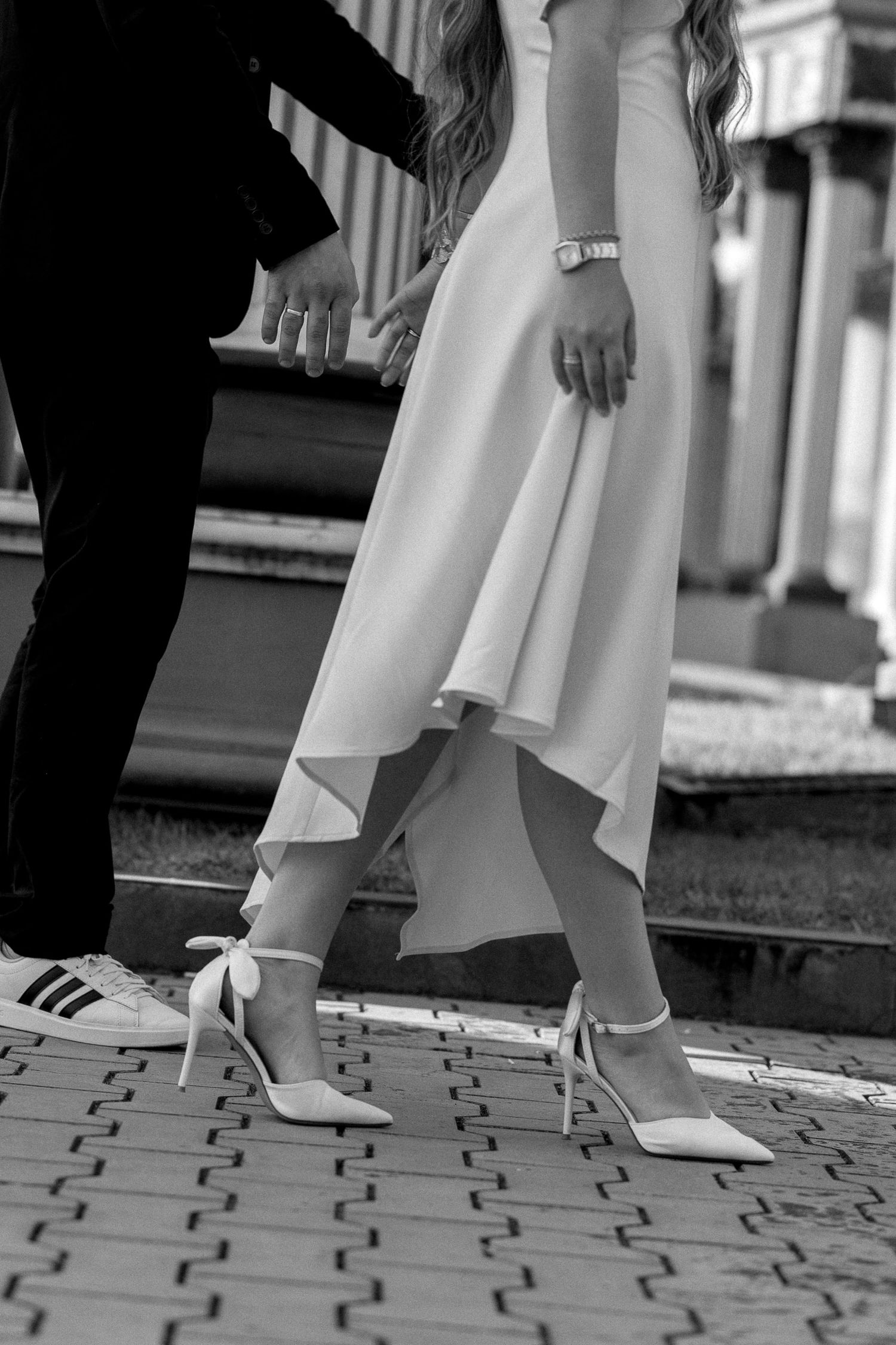 Black and white photo of bride in white dress and heels holding hands with groom in suit on cobblestone steps.