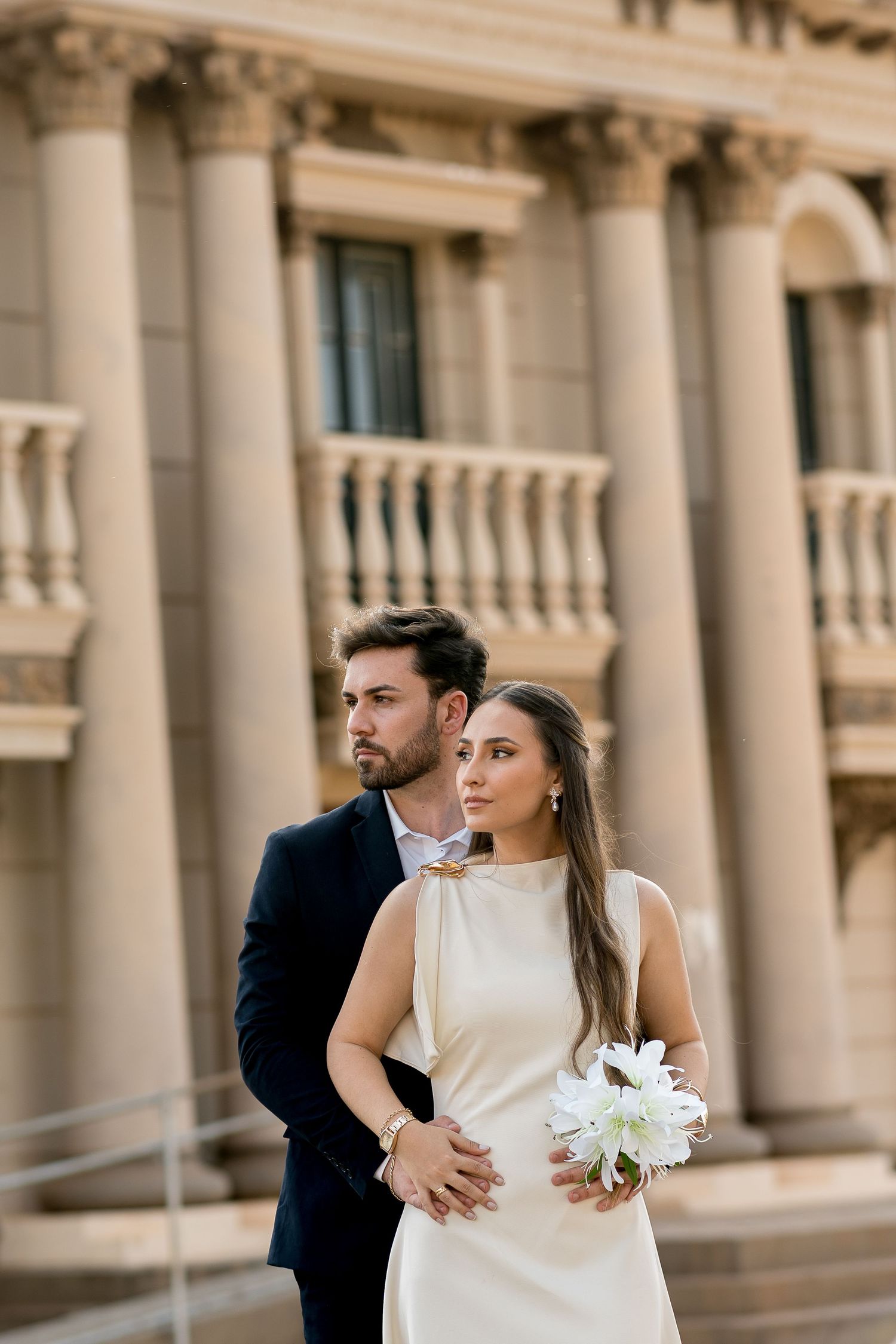 Elegant couple in wedding attire posing before a classical stone building with columns, bride holding white lilies.
