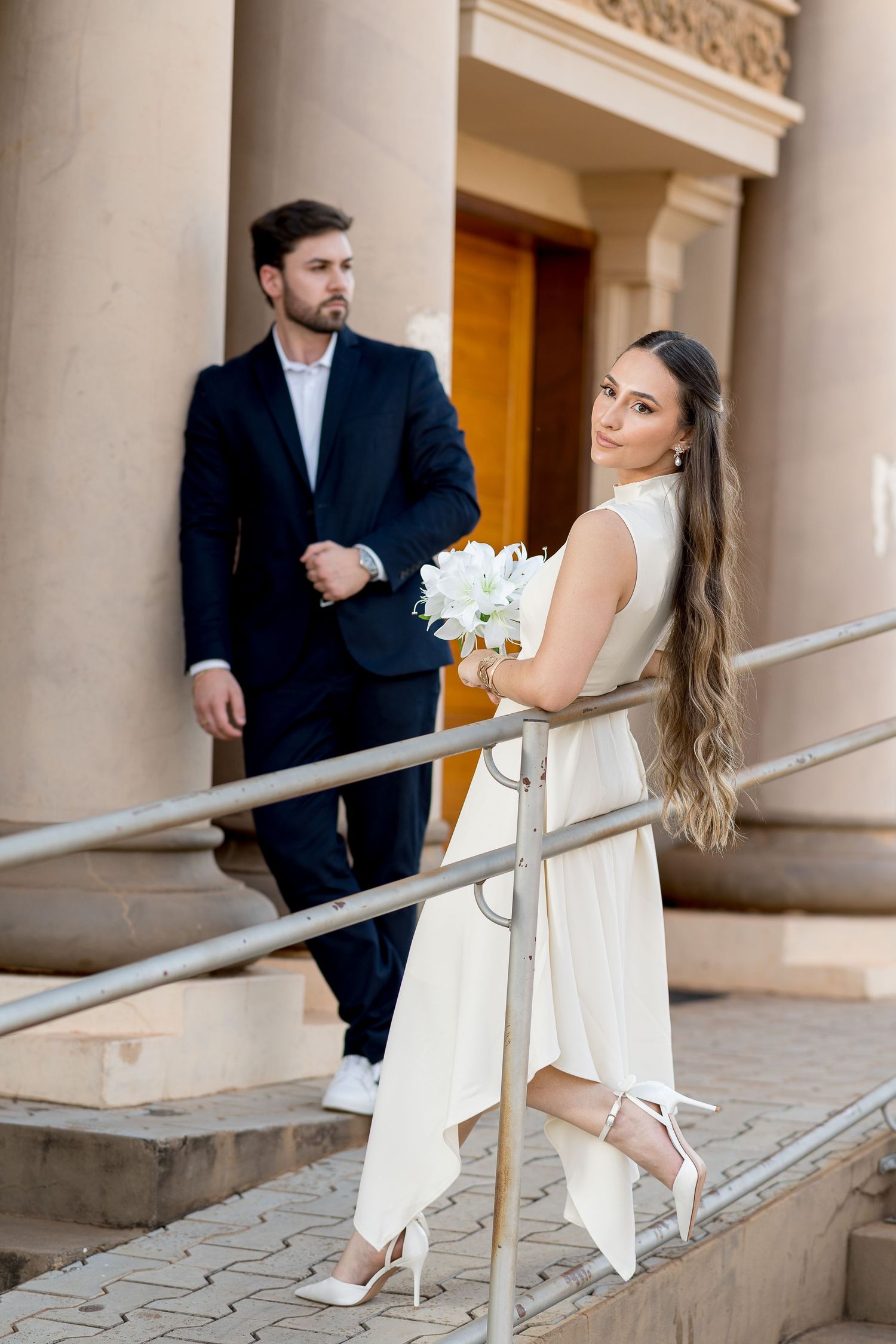 Elegant bride in white dress holds bouquet on steps while groom in dark suit stands nearby at a classic building.