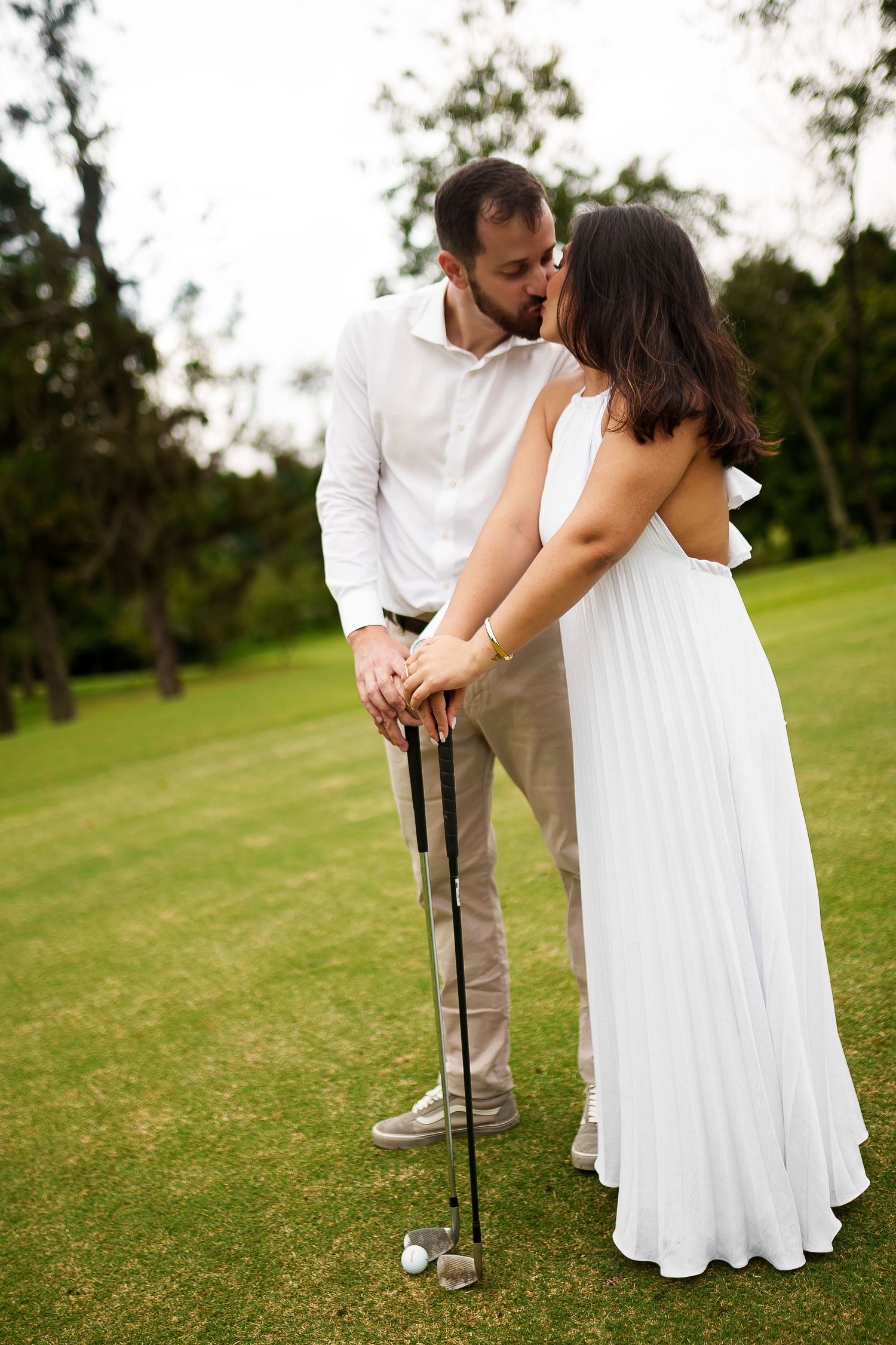 Couple sharing a romantic kiss on a golf course, dressed in white and beige, holding golf clubs on green grass.