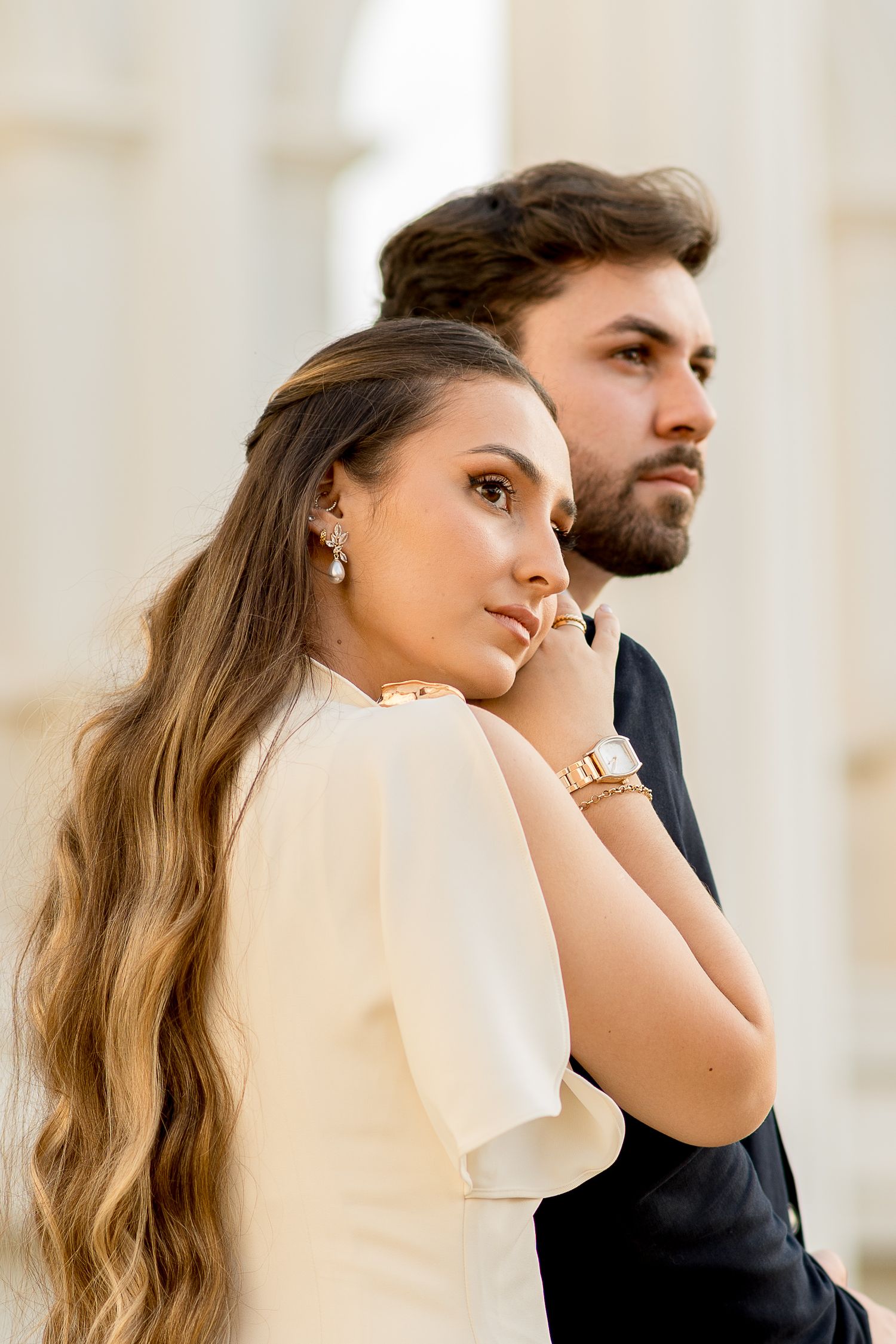 Elegant couple in formal attire posing outdoors, woman in cream dress with gold jewelry, man in black suit behind her.