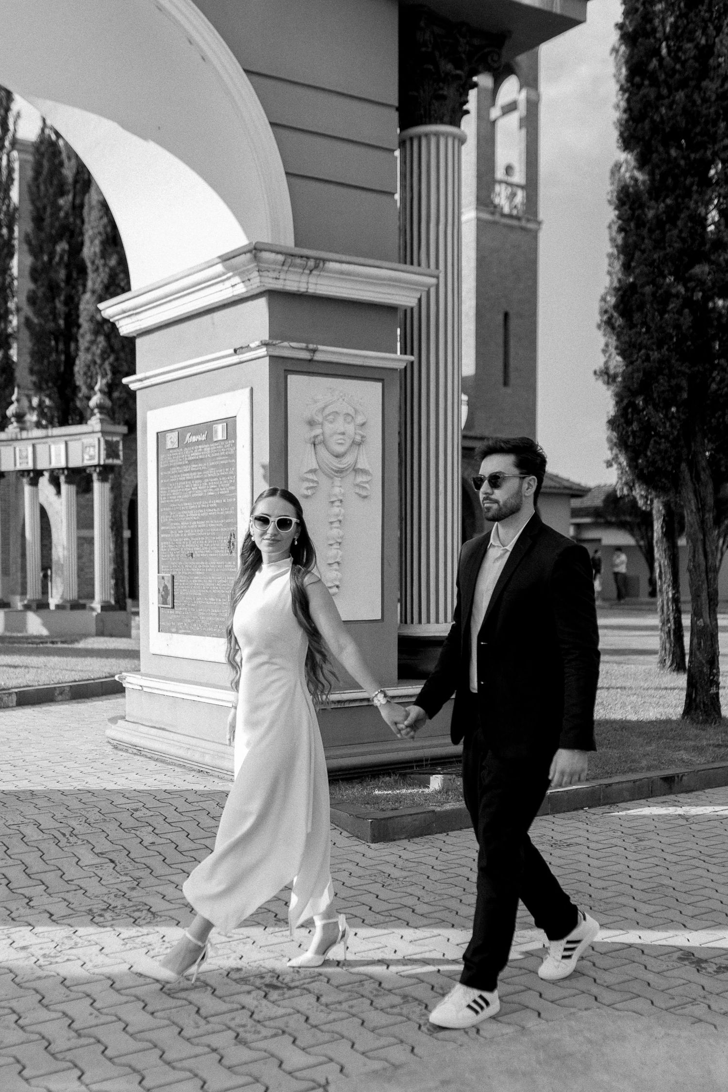 Black and white photo of an elegant couple holding hands, woman in white dress and man in suit, walking outdoors near ornate architecture.