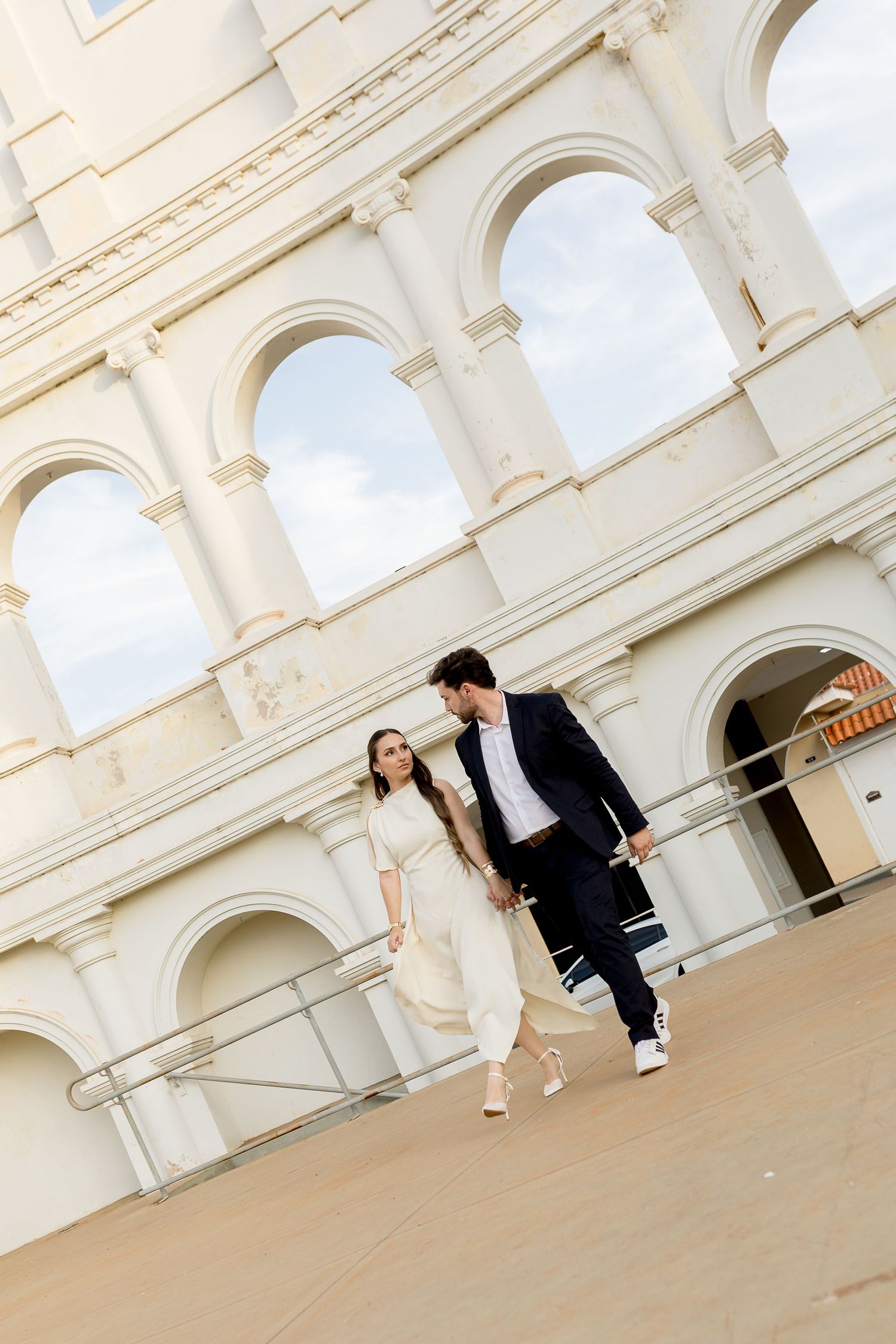 Couple holding hands walking down steps in front of a grand white Roman Colosseum-style building.