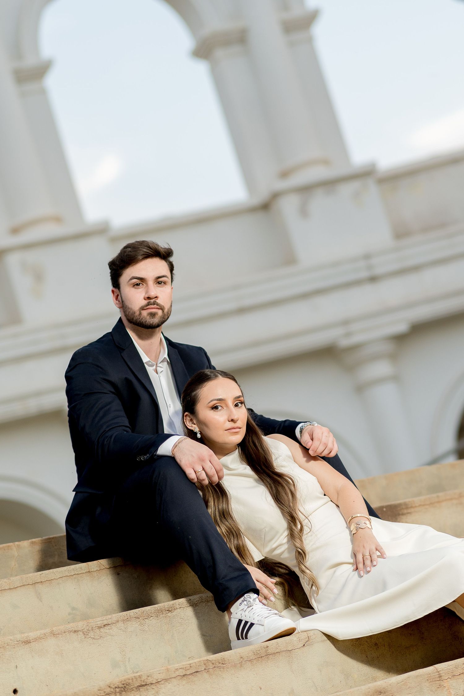 Elegant couple sitting on stone steps near classical arches, man in dark suit, woman in white dress and Adidas sneakers.