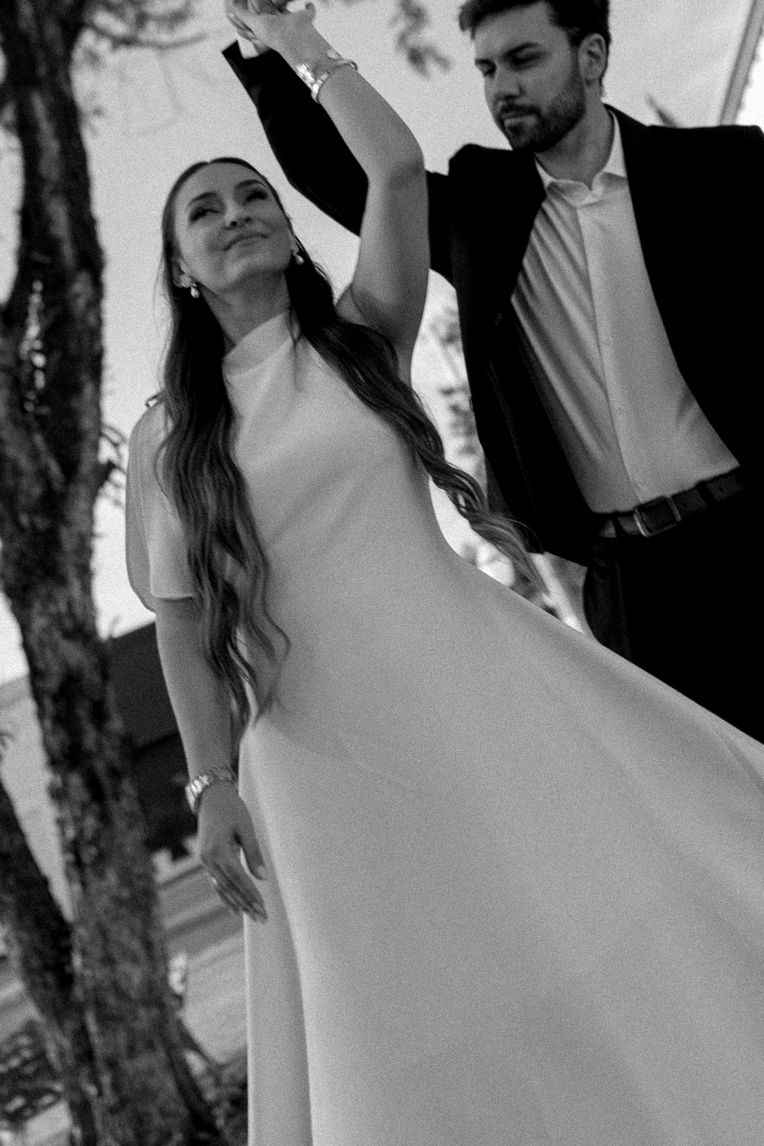 Black and white photo of a bride in white gown dancing with groom in suit outdoors near a tree.