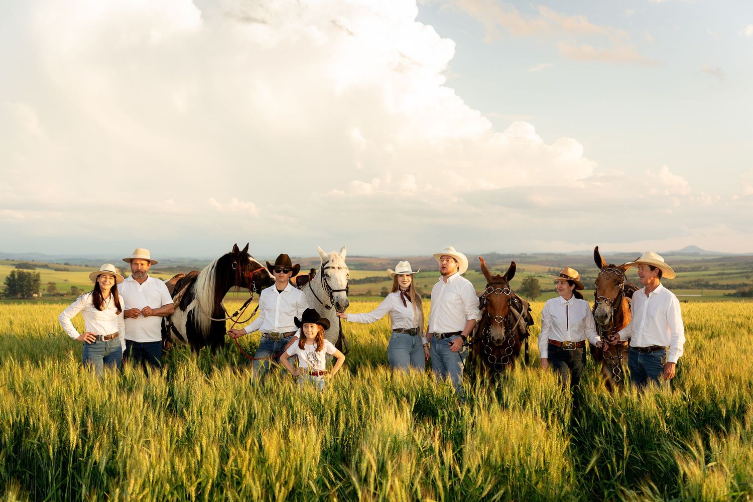 A cowboy family poses with horses in a lush green field under dramatic clouds in a scenic rural landscape.