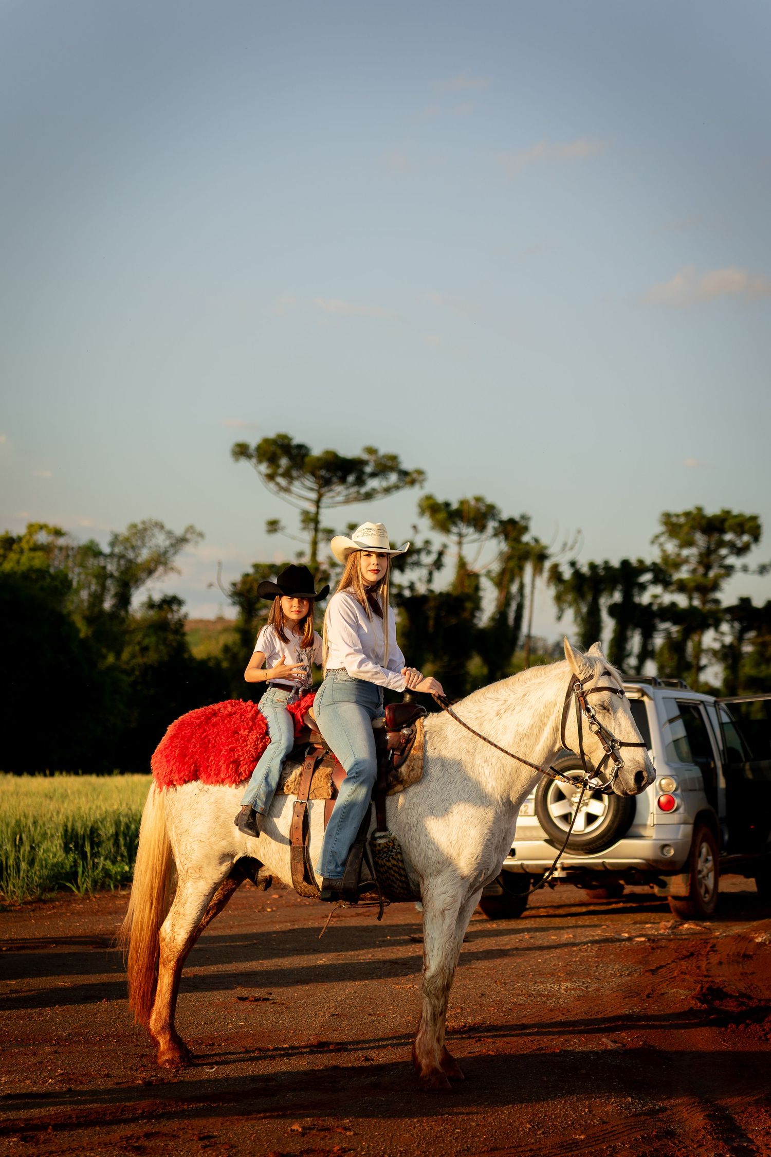 Two women riding a white horse on a dirt path at golden hour, with green fields and trees in the background.