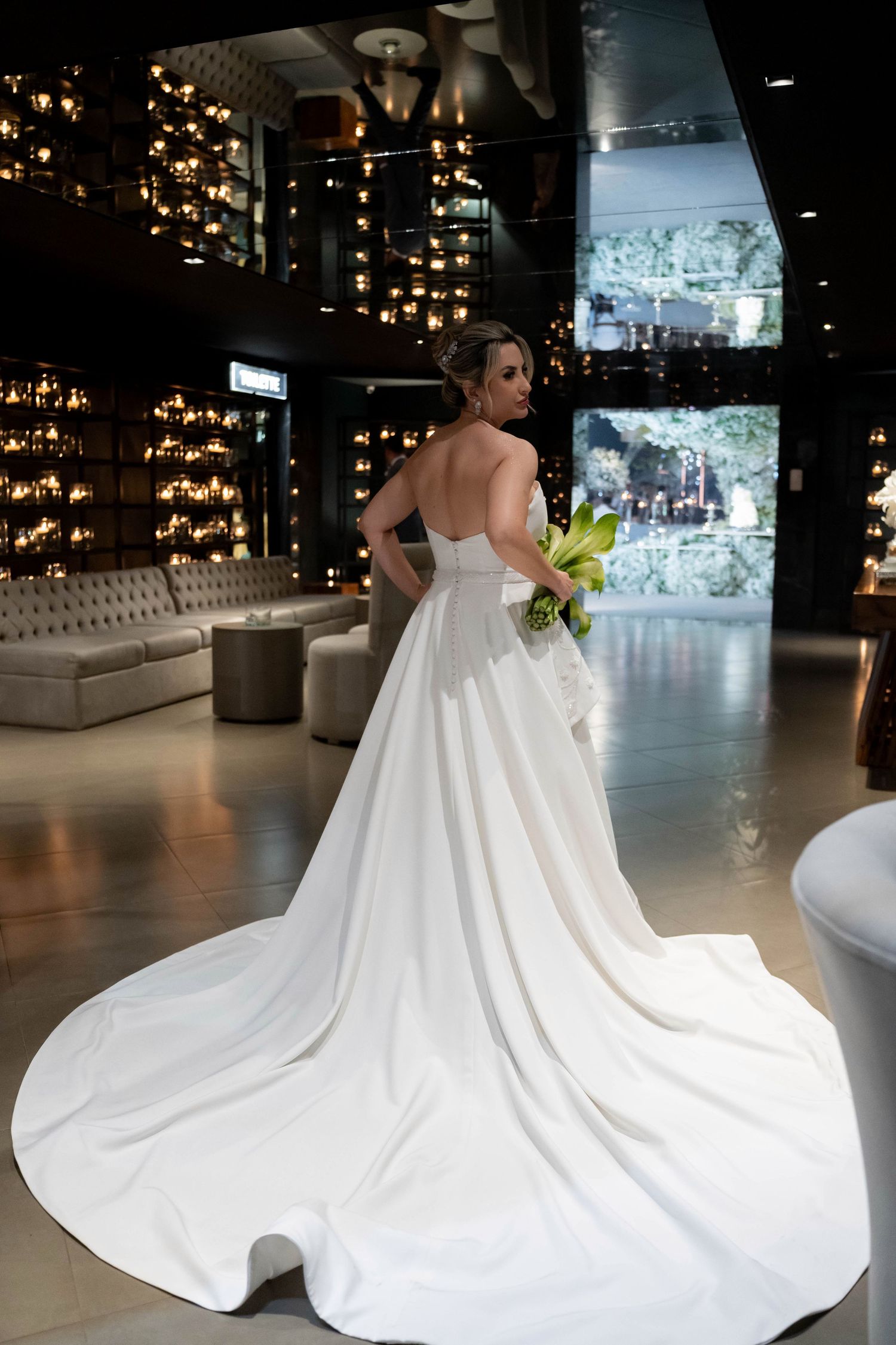 Bride in elegant strapless white ball gown with long train, holding green bouquet, posing in luxurious venue.