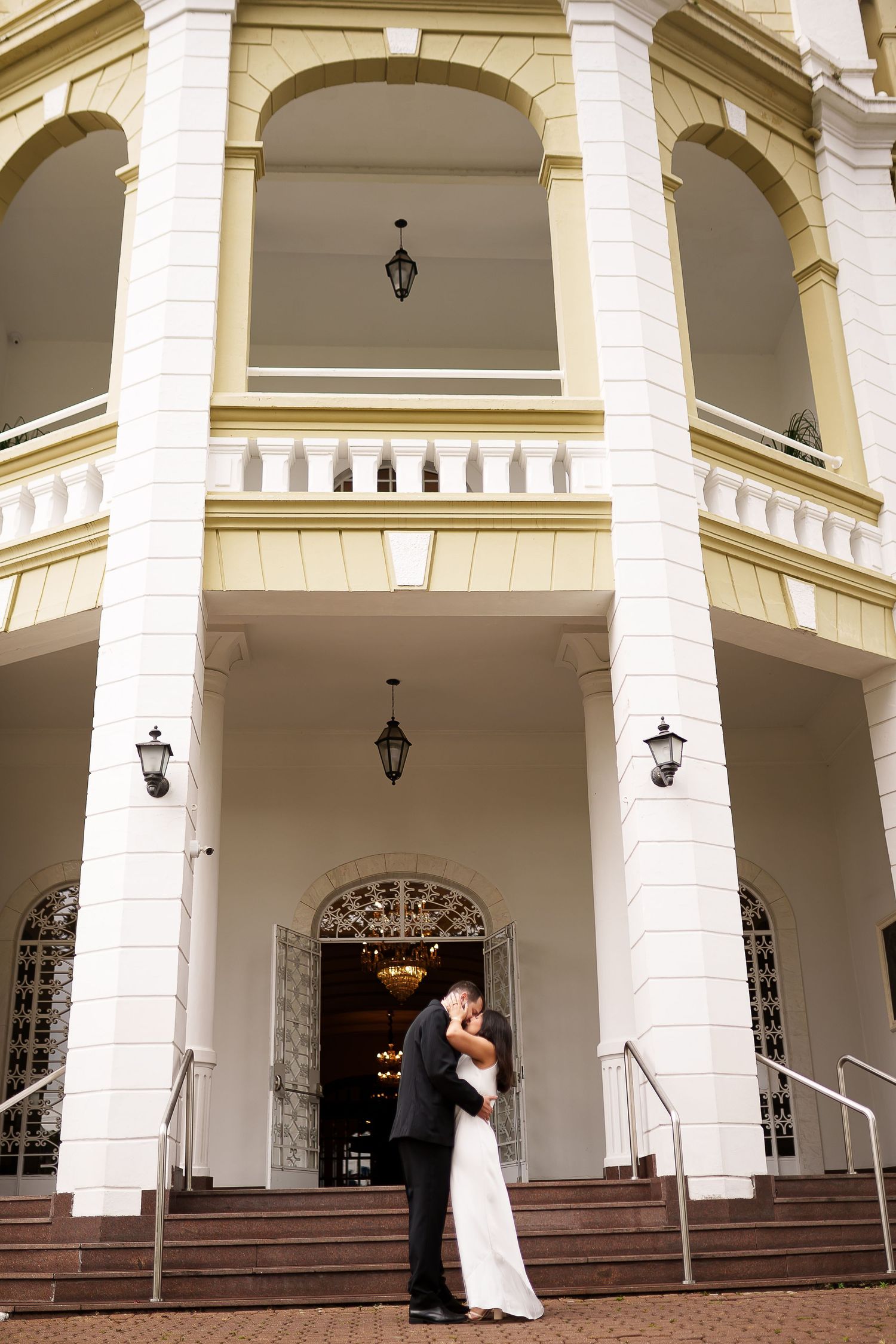 Bride and groom share a romantic kiss in front of an elegant white colonial building with grand columns.