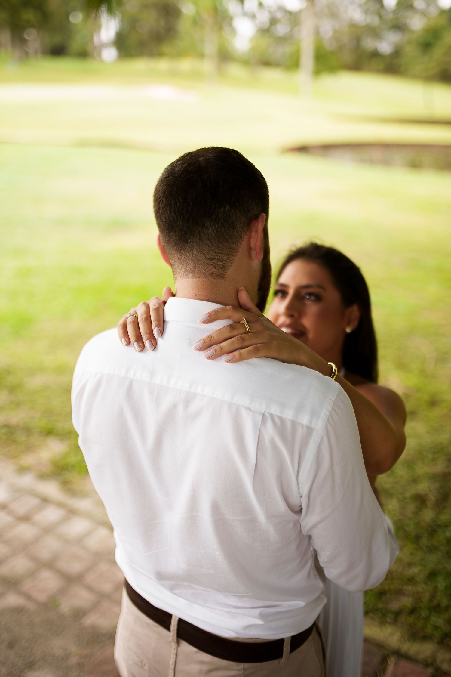 A couple embraces outdoors, woman with dark hair rests her hand on man's shoulder wearing a gold ring, green park background.