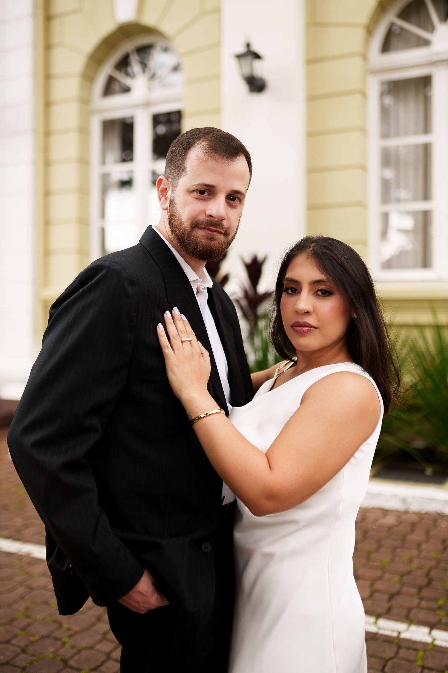 Elegant couple posing outdoors, man in black suit and woman in white dress, embracing in front of a classic building.