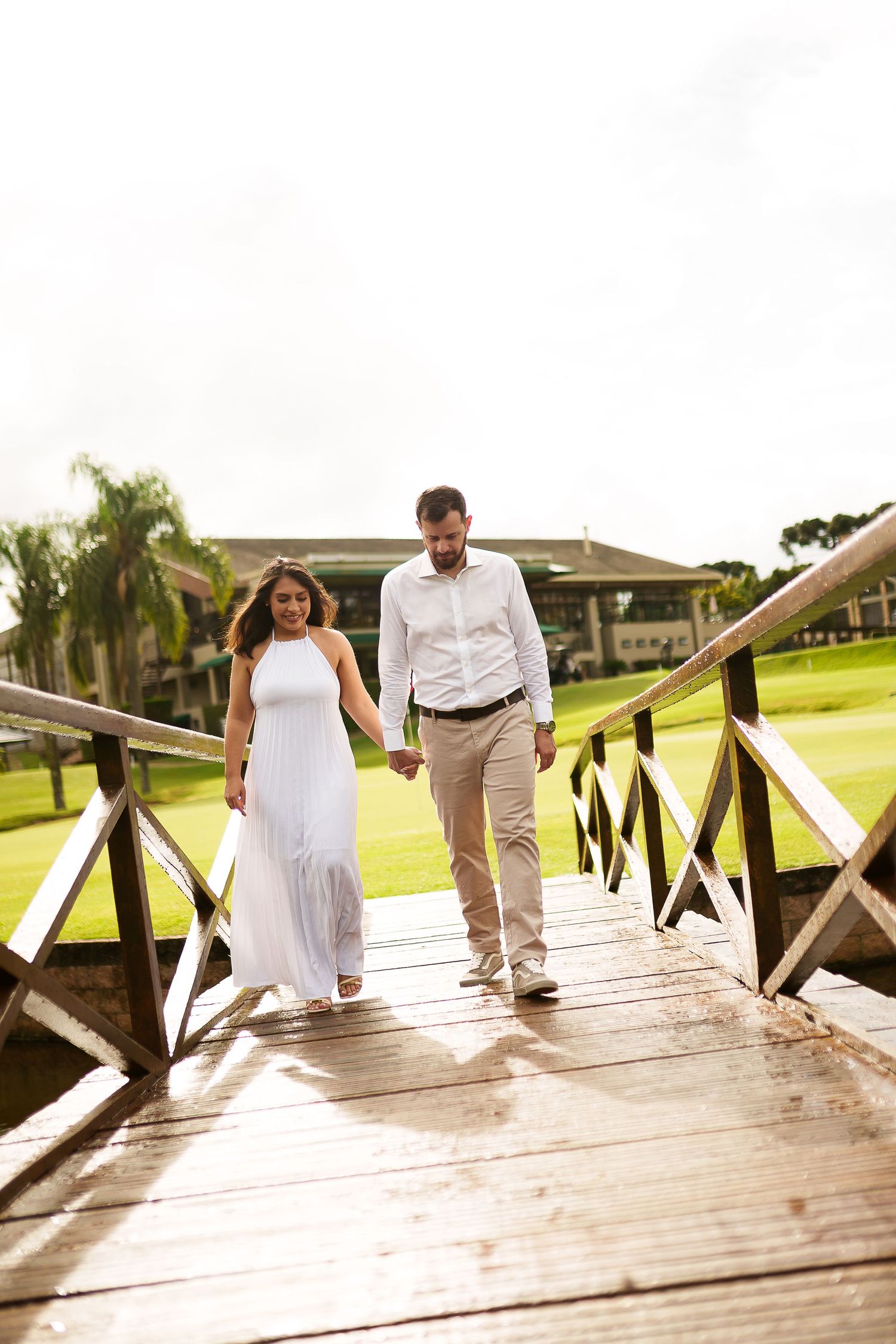 Couple holding hands walking across a wooden bridge, woman in white dress, man in white shirt, green golf course background.