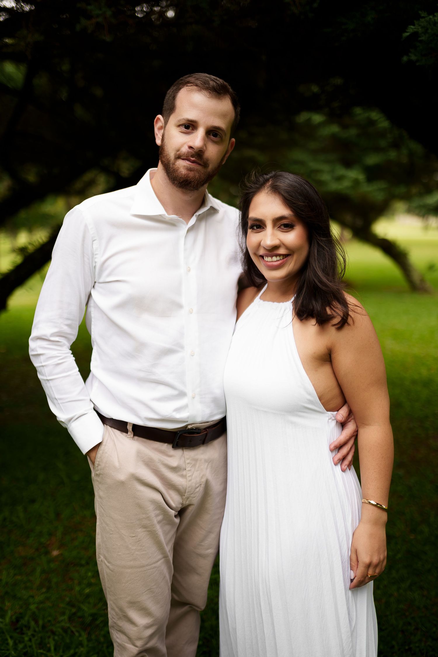 Couple in white outfits posing outdoors in a lush green park setting for an engagement photo.