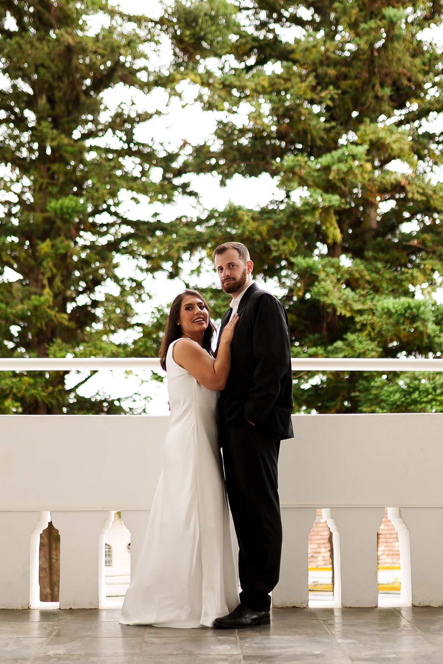 Bride and groom posing on outdoor terrace with lush green trees, bride in white gown, groom in black suit.