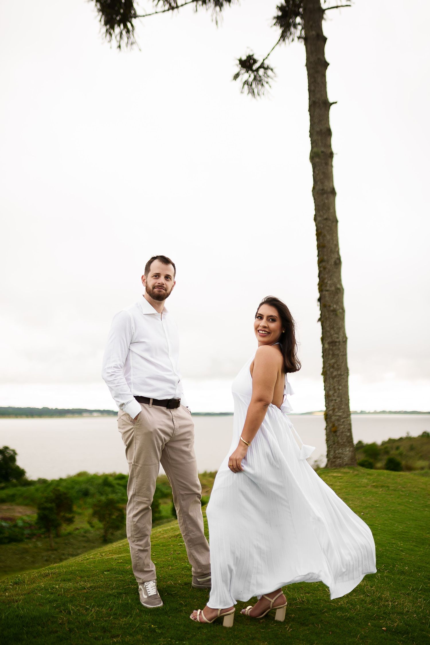 Couple in white attire standing on green lawn near tall tree with lake view, woman in flowing dress smiling over shoulder.