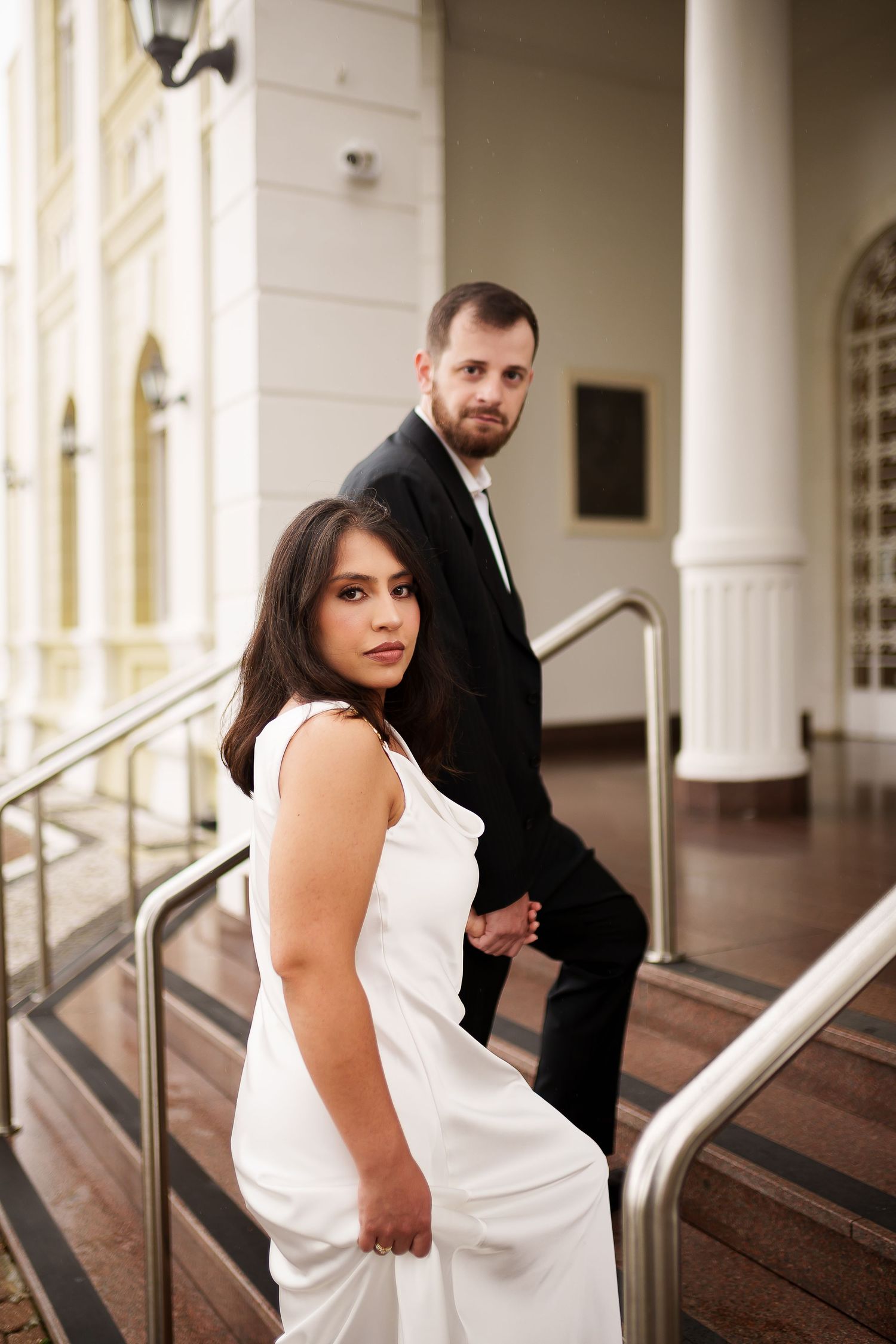 Elegant couple in formal attire posing on grand building steps, woman in white gown and man in black suit.