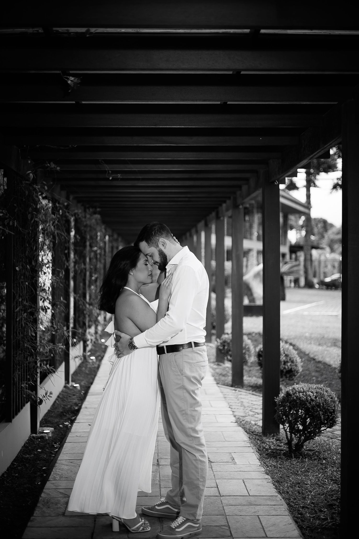 Black and white photo of a romantic couple embracing and kissing under a pergola walkway with lush greenery.