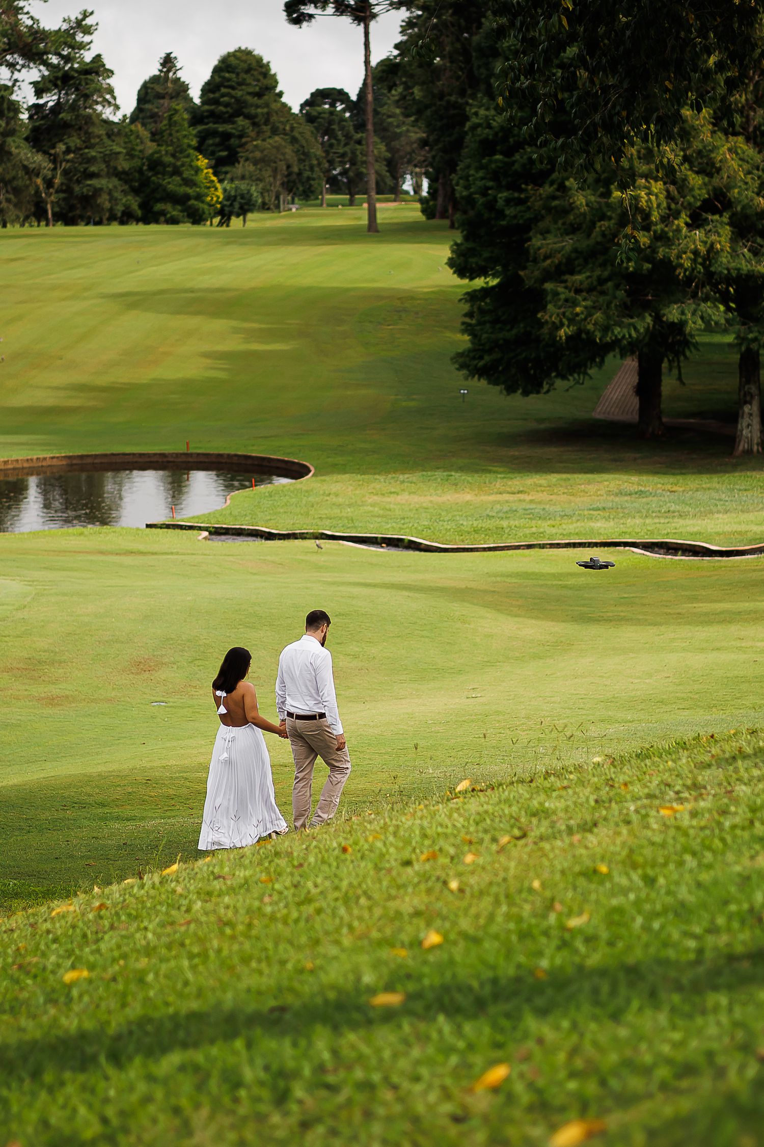 Couple in white dress and khakis walking hand-in-hand on lush green golf course with pond and trees.