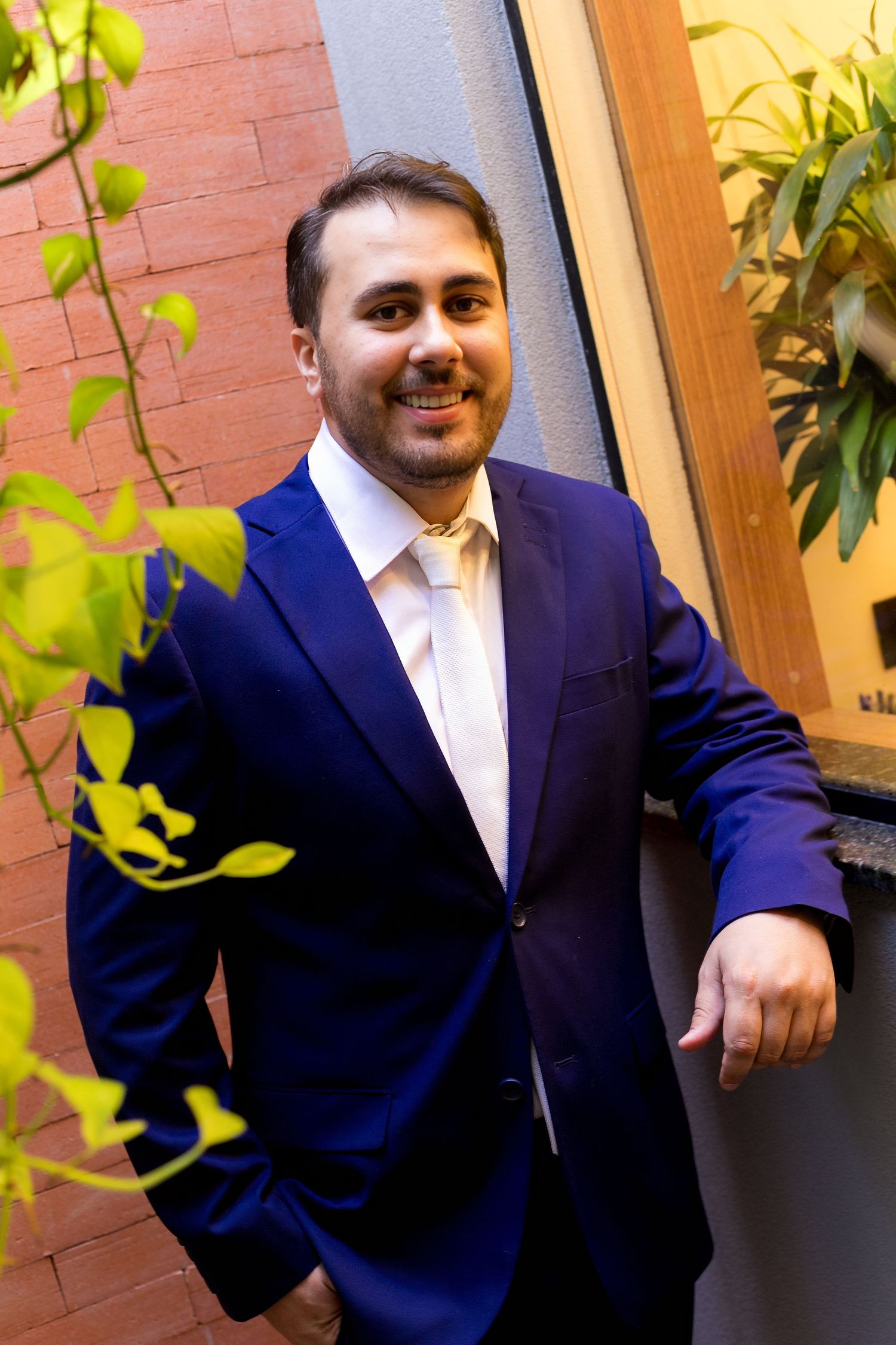 Smiling man in blue suit and gold tie posing outdoors near brick wall and green plants.