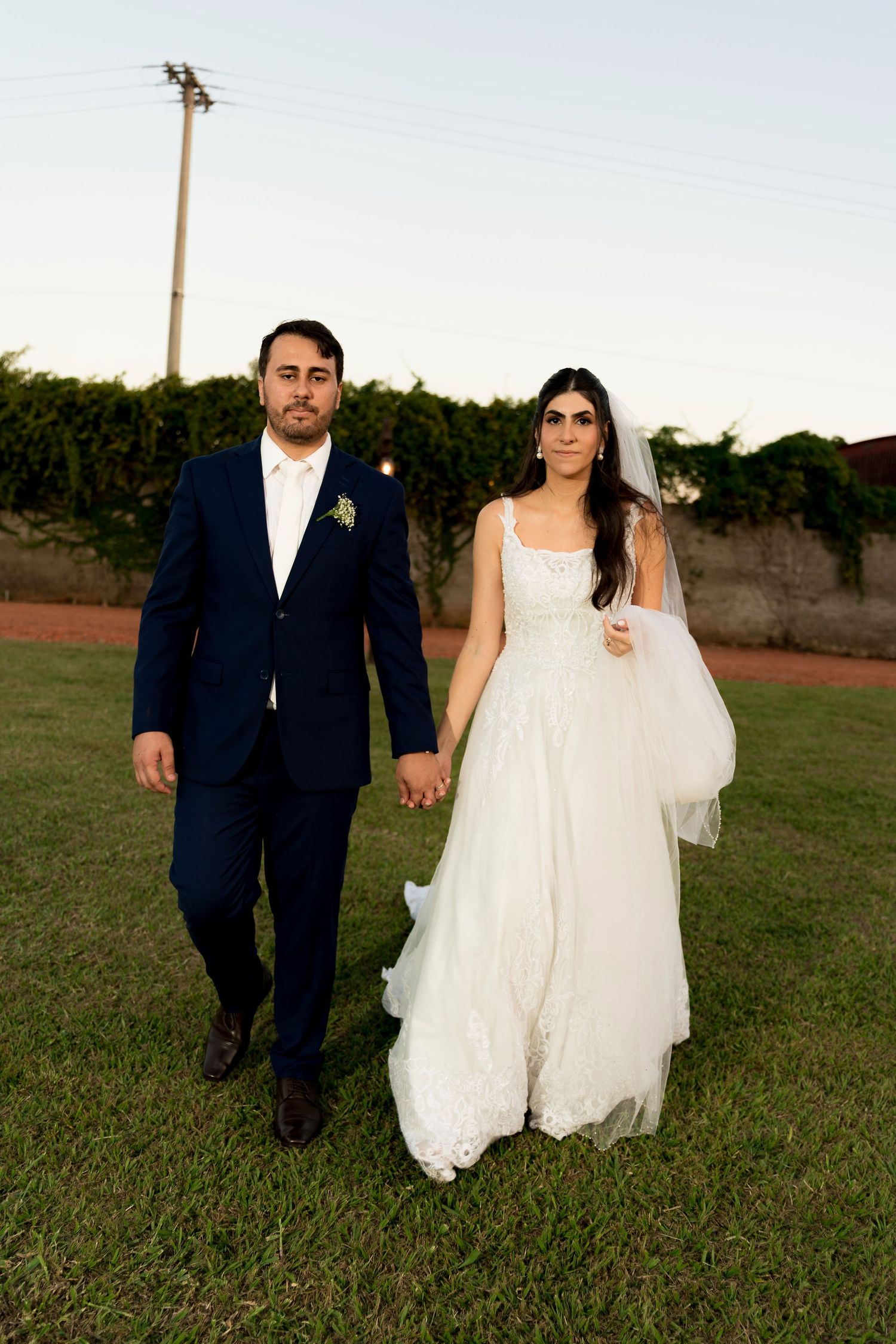 A bride in a white gown and groom in a navy suit hold hands walking on grass at their outdoor wedding ceremony.