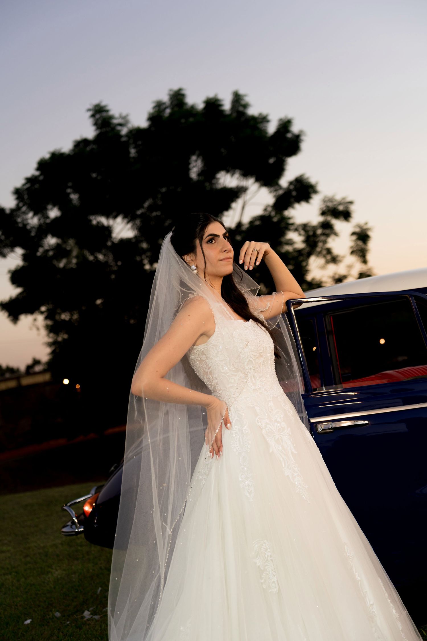 Bride in white lace ball gown and veil poses beside vintage blue car at dusk with trees in background.