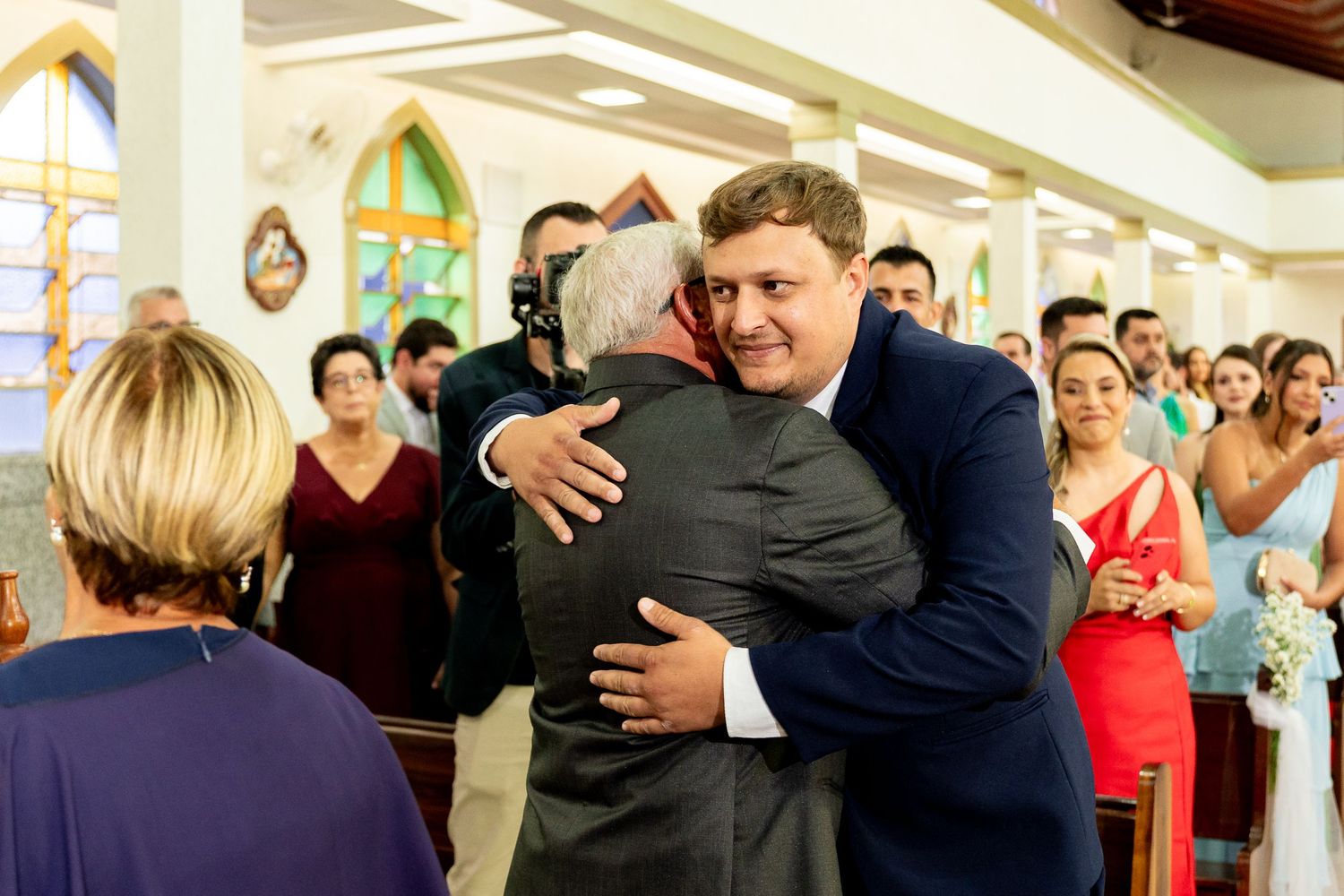 Two men share a warm hug at a wedding ceremony inside a decorated church with guests in the background.