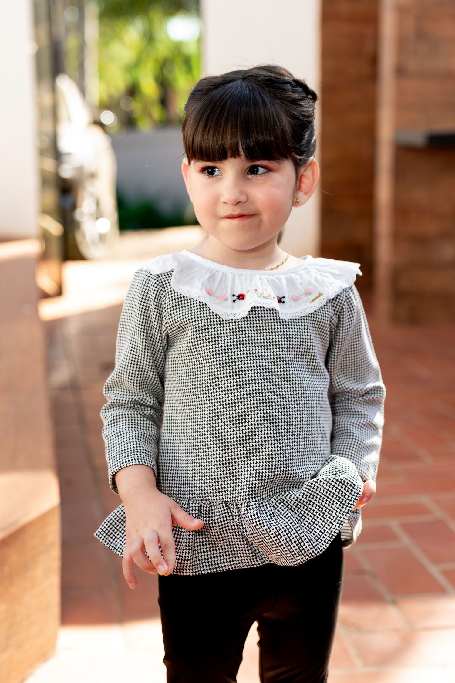 Young girl in houndstooth top with white lace collar, posing outdoors on terracotta tiles.