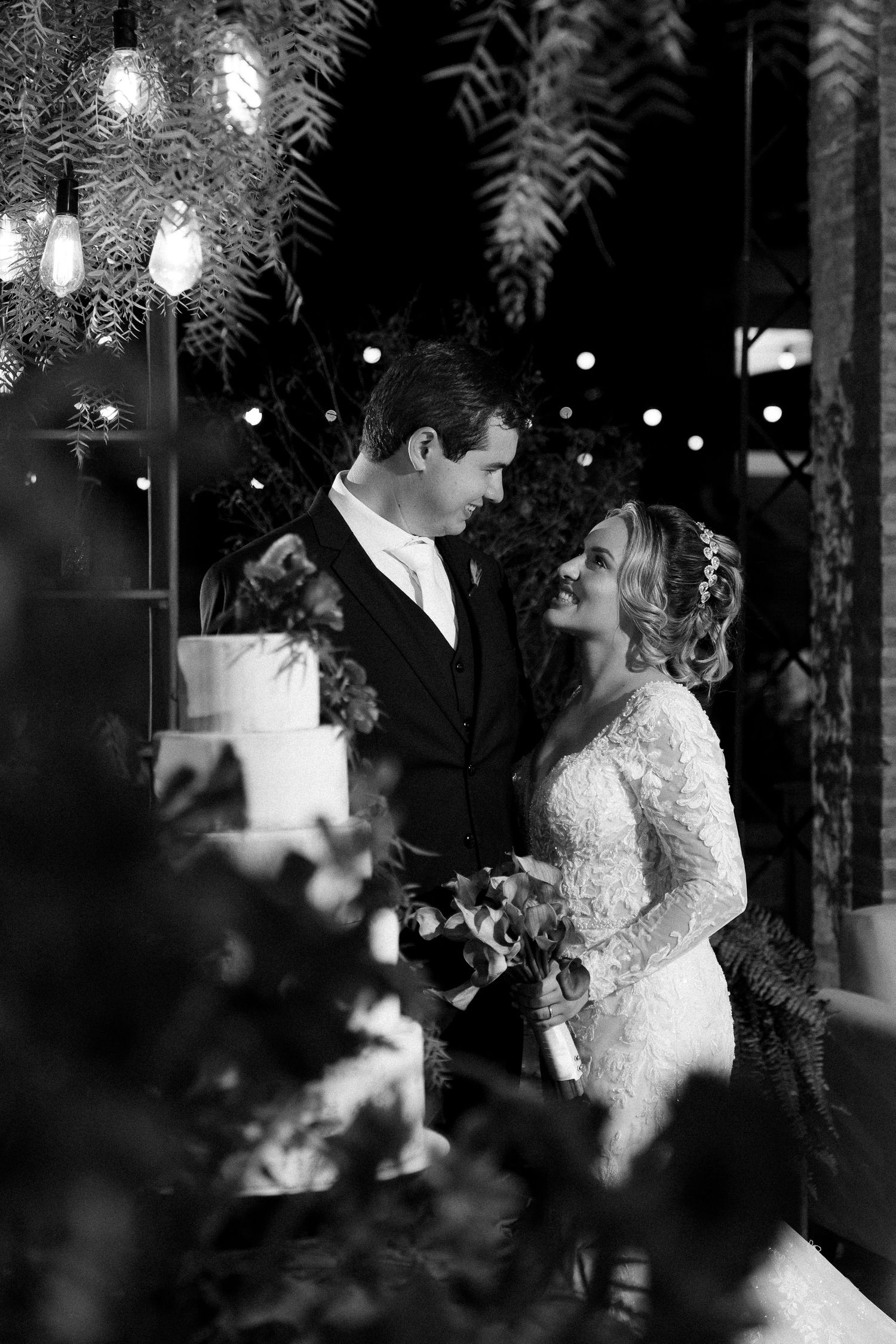 Black and white wedding photo of bride and groom sharing a romantic moment beside tiered cake amid lush greenery.