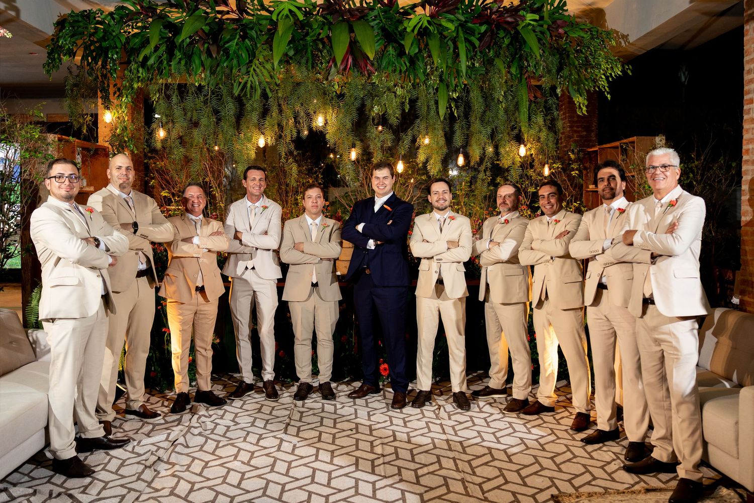 Groom in navy suit poses with groomsmen in tan suits under lush greenery at elegant wedding venue.