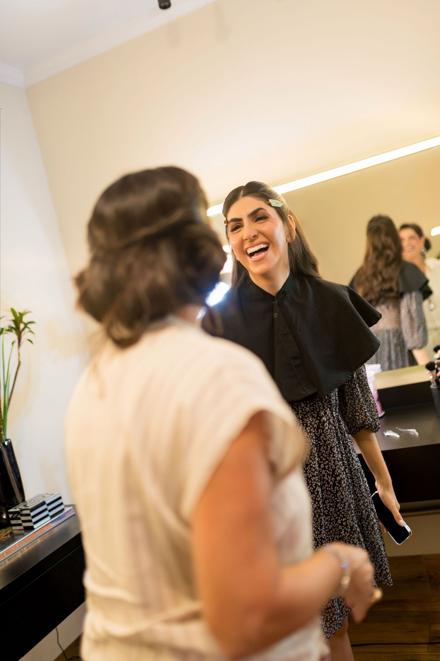 Two women laughing together in a beauty salon, one wearing a black dress with a cape collar near a lit mirror.