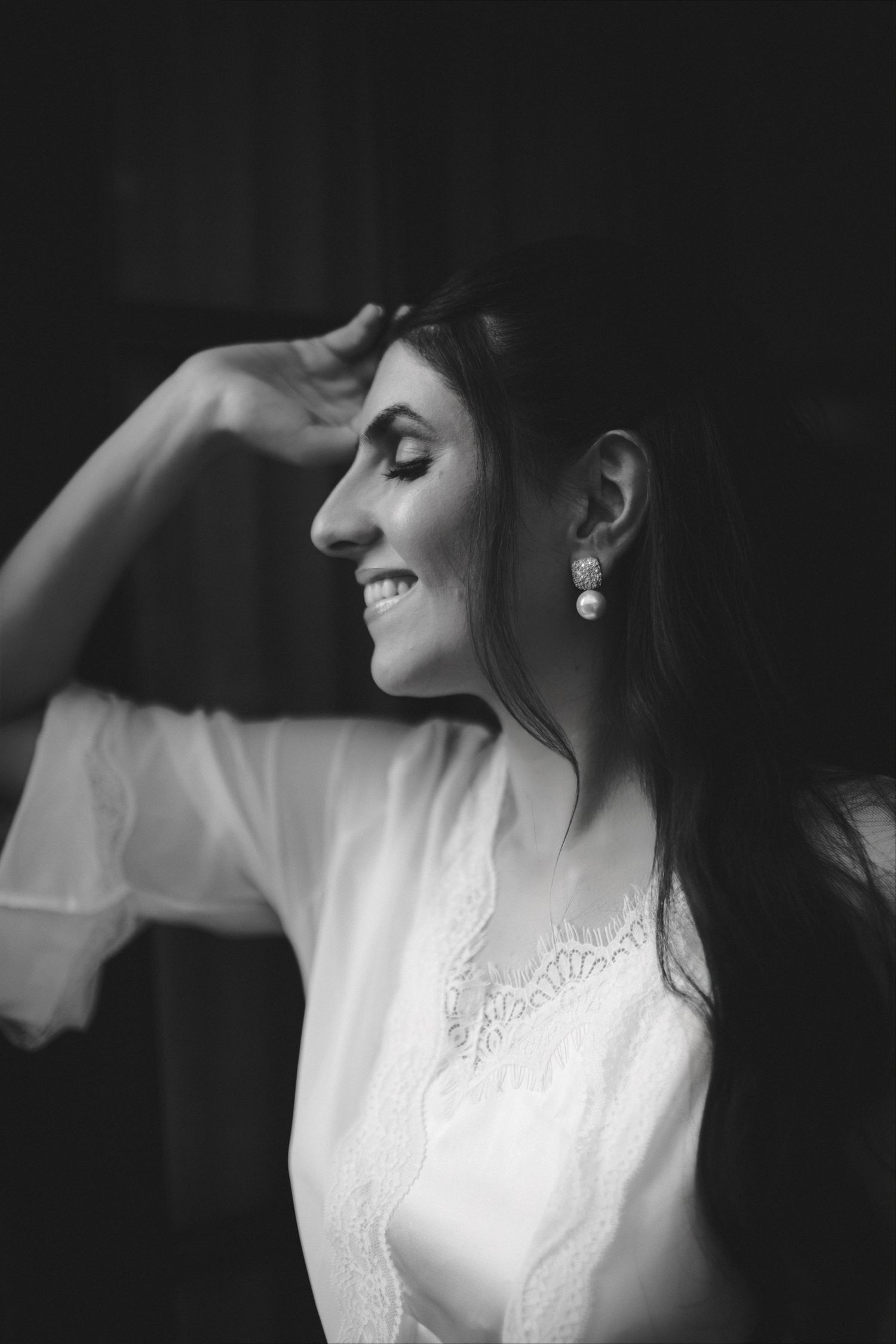 Black and white portrait of a smiling bride in white lace robe wearing pearl earrings, hand raised to her head.
