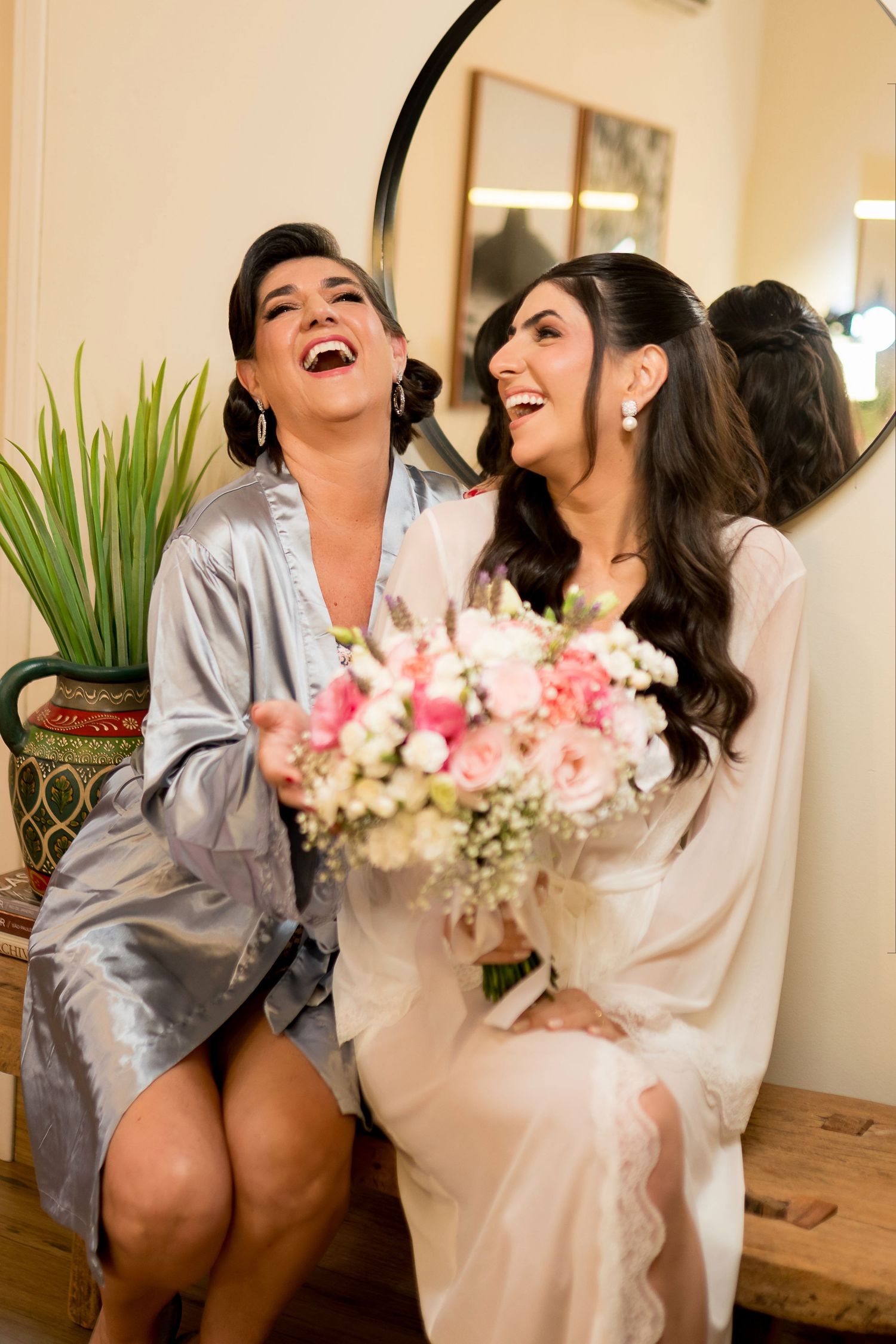 Two women laughing joyfully, bride in white robe holding pink bouquet with bridesmaid in silver robe.