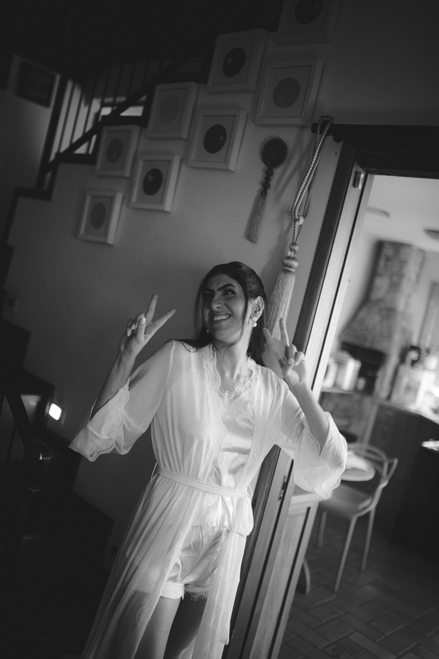 Black and white photo of a smiling woman in a white robe making peace signs inside an elegant home.