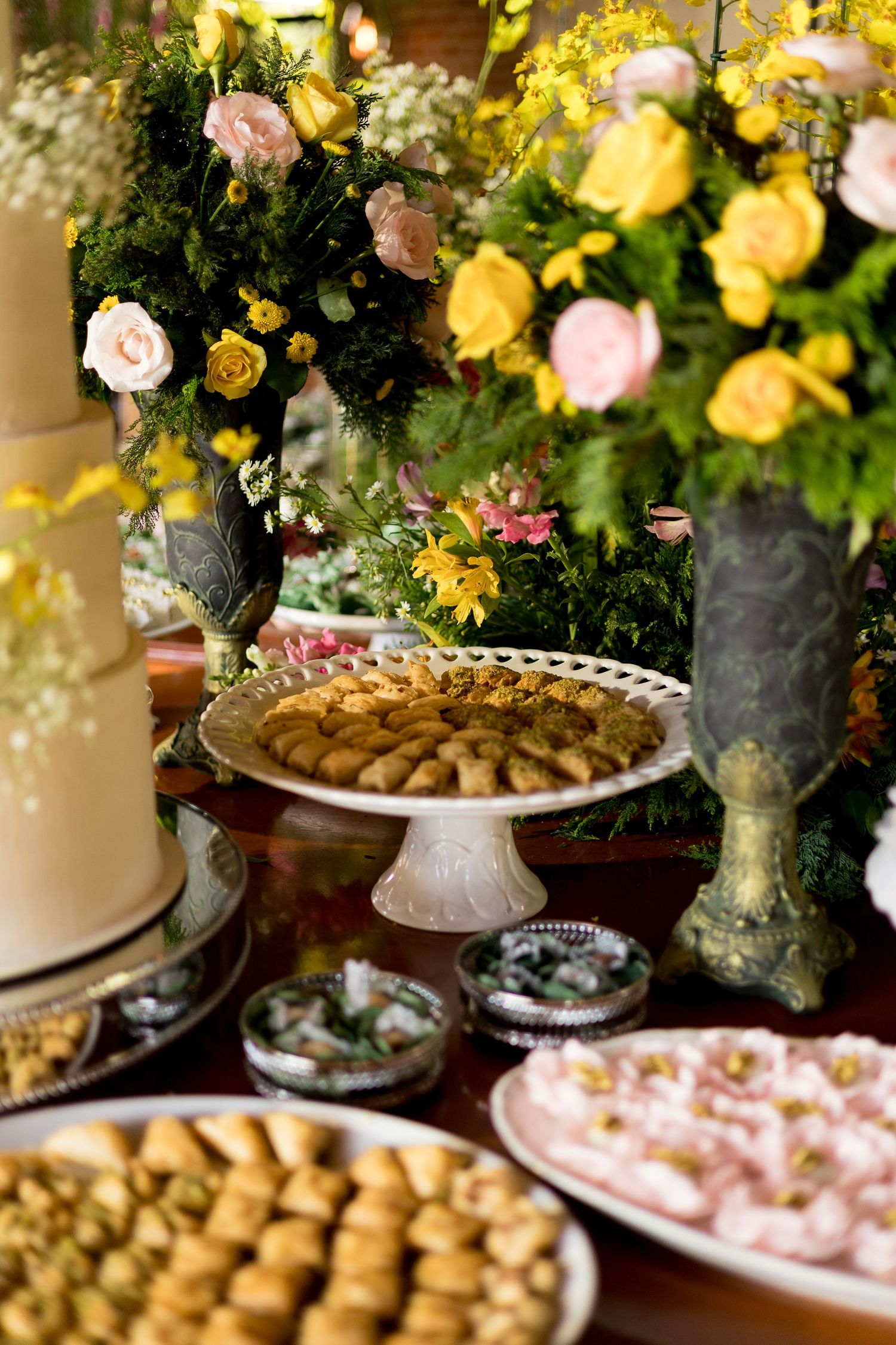 Elegant dessert table with assorted pastries, a white cake, and lush floral arrangements featuring yellow and pink roses.