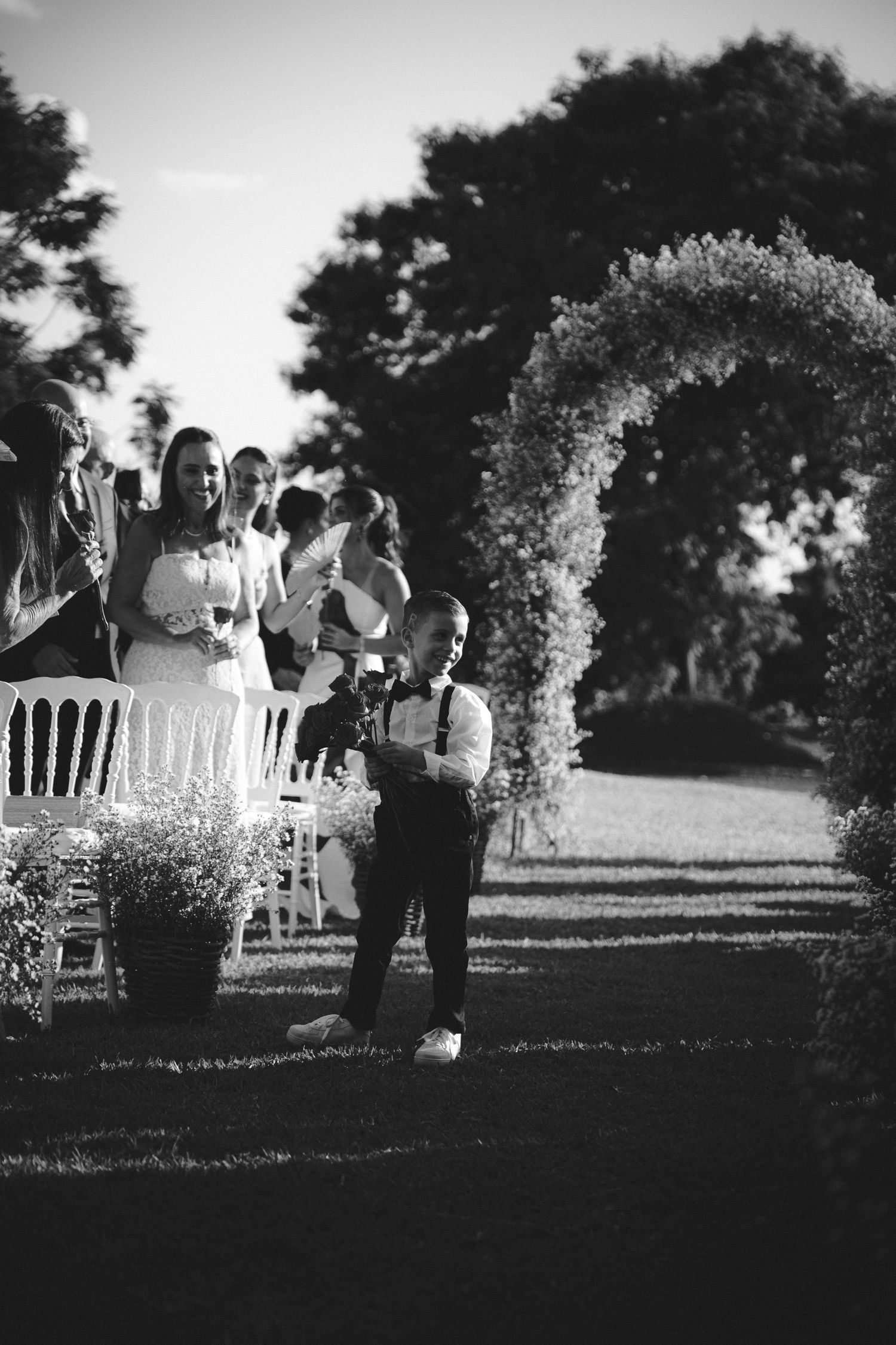 Black and white outdoor wedding ceremony with bride, guests seated in white chairs, and a young boy near a floral arch.