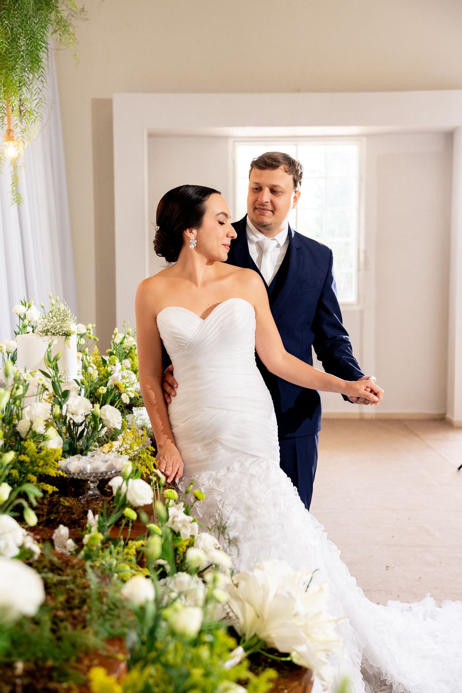 Bride in white strapless mermaid gown and groom in navy suit walk together past lush white floral arrangements.