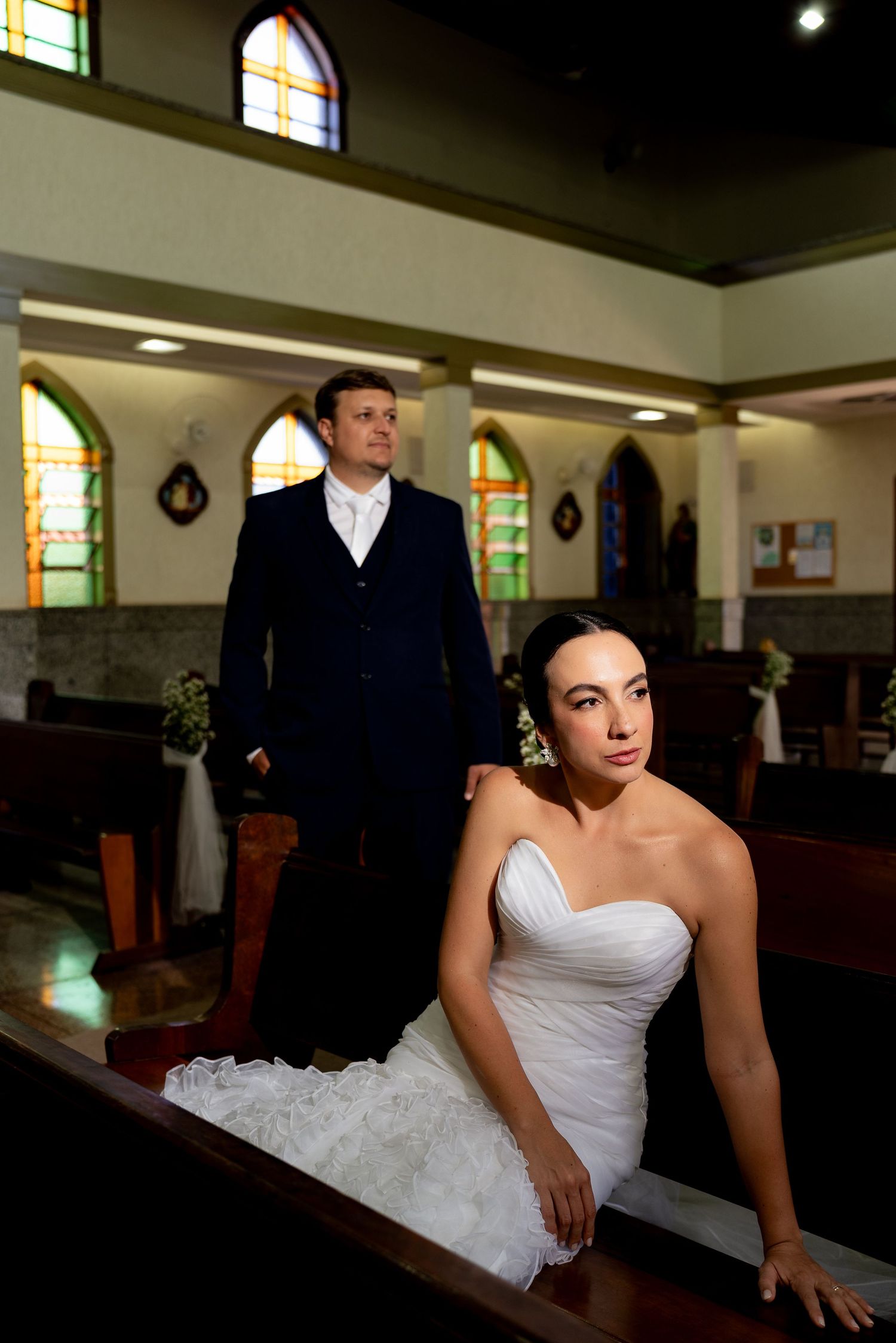 Bride in white strapless gown sits on church pew while groom in navy suit stands behind her in a sunlit chapel.