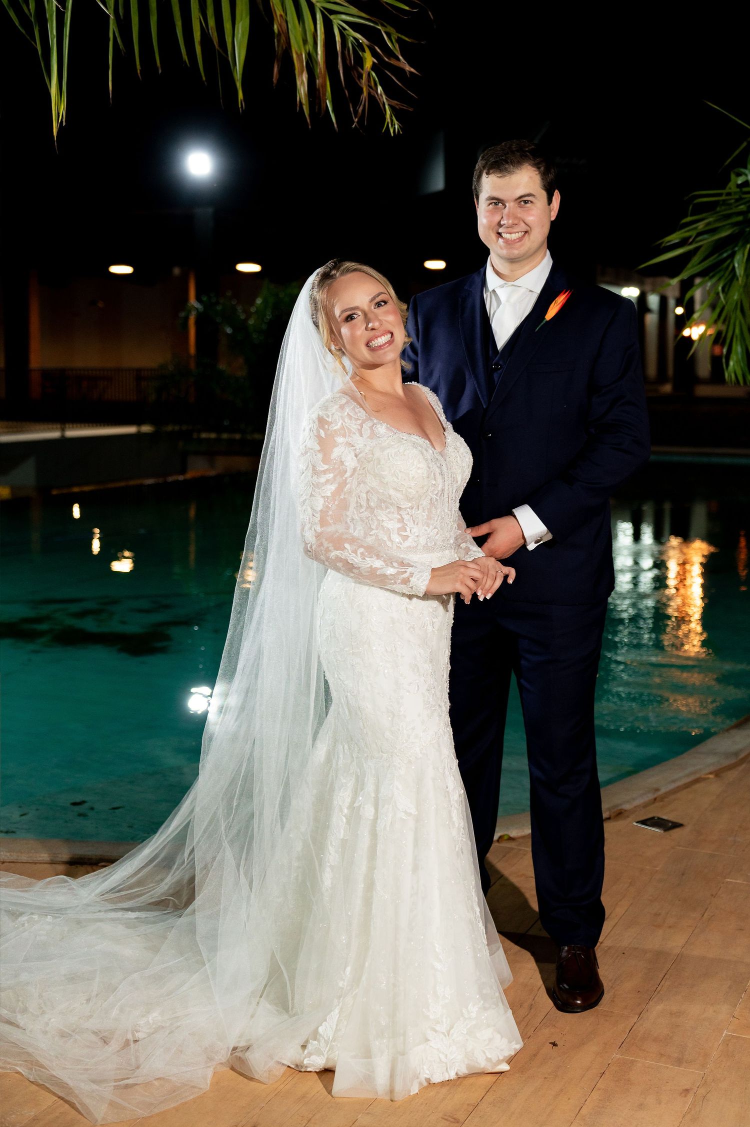 Bride in lace gown with veil and groom in navy suit pose by illuminated pool at night wedding venue.