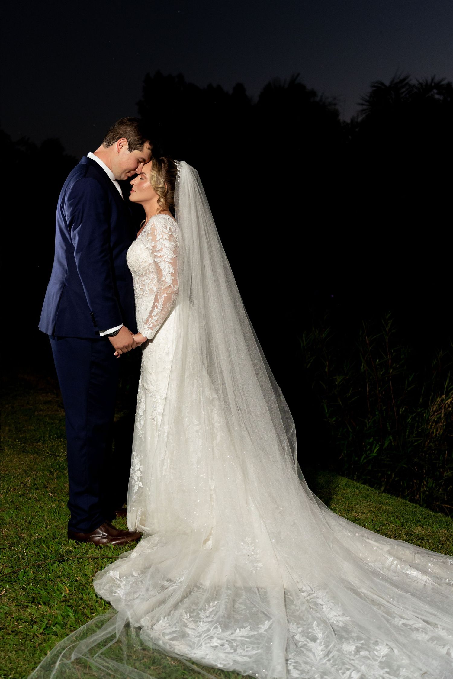 Bride and groom share a romantic moment outdoors at night, bride wearing lace gown with long veil.