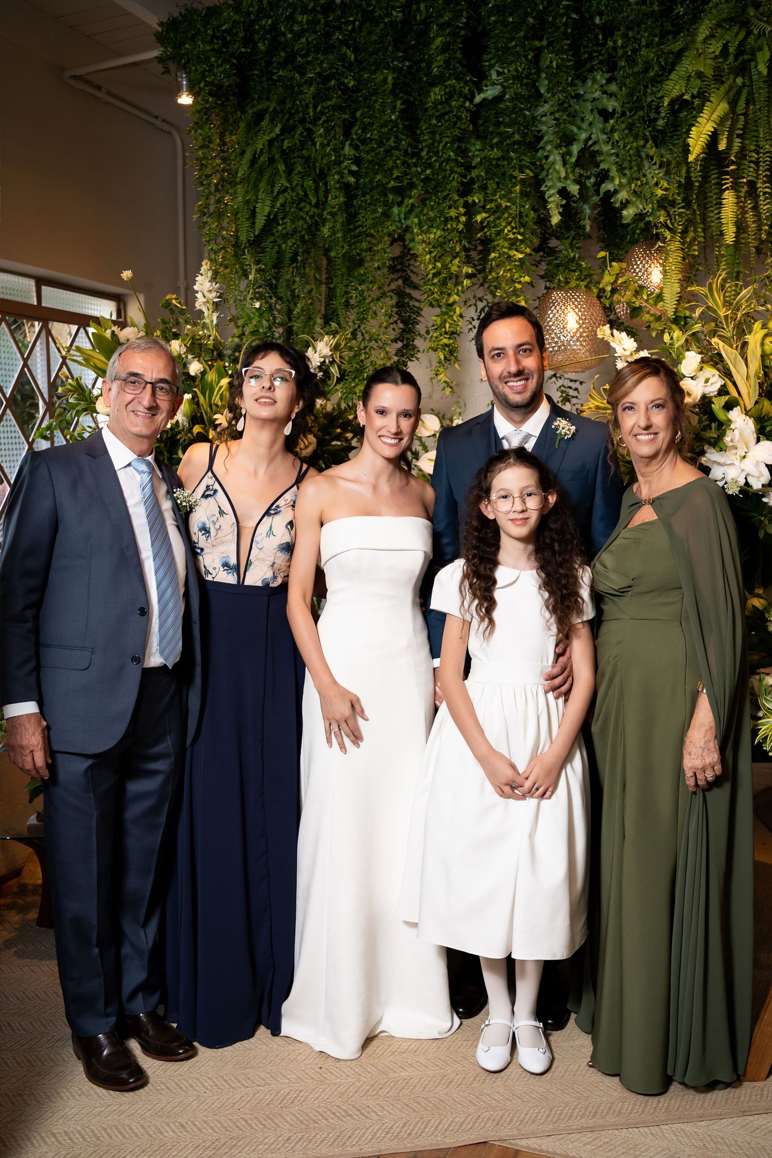 Wedding party poses with bride in white gown, groom in navy suit, and family in formal attire before lush greenery backdrop.