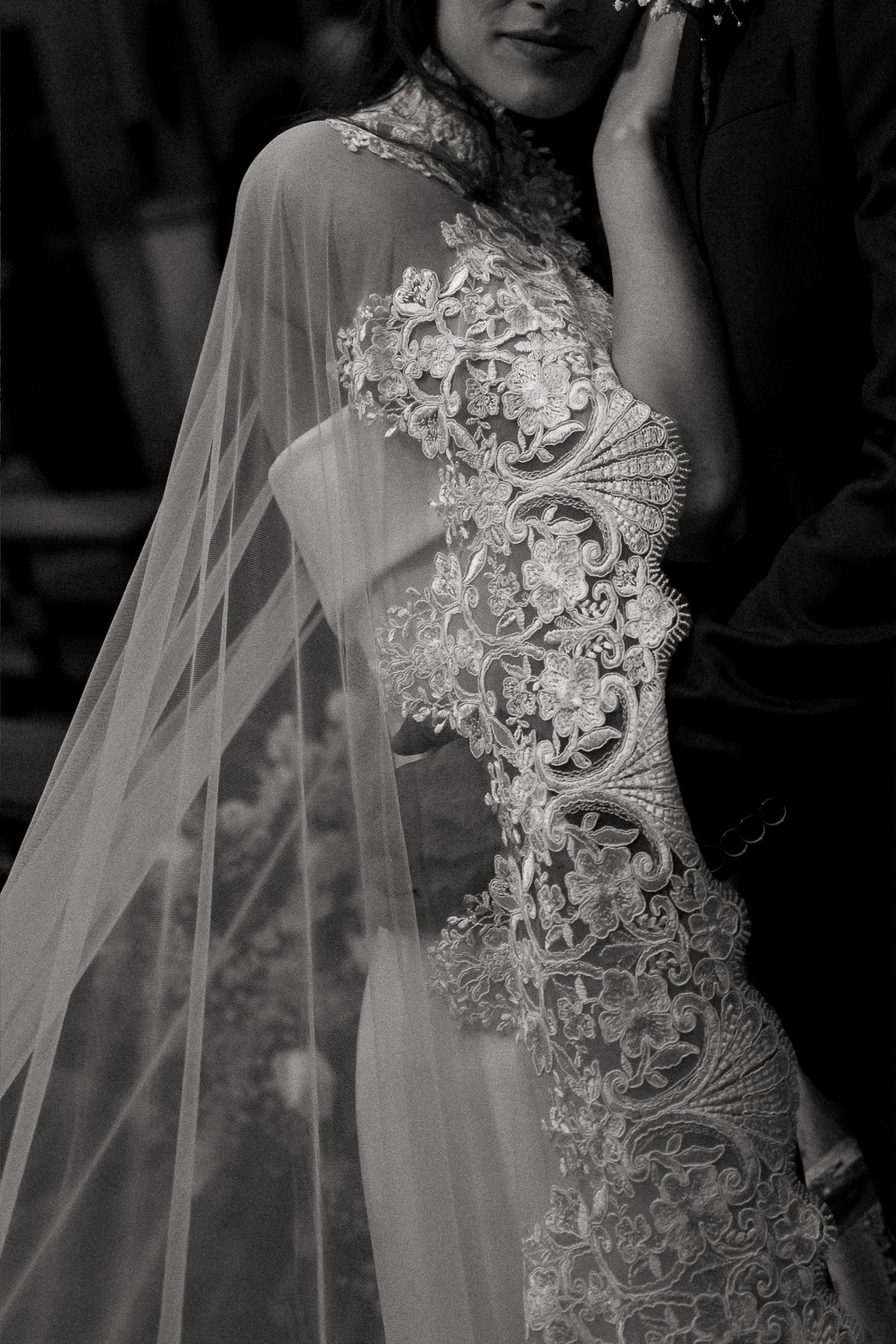 Black and white photo of a bride in an intricate lace wedding gown with a flowing veil, embraced by her groom.